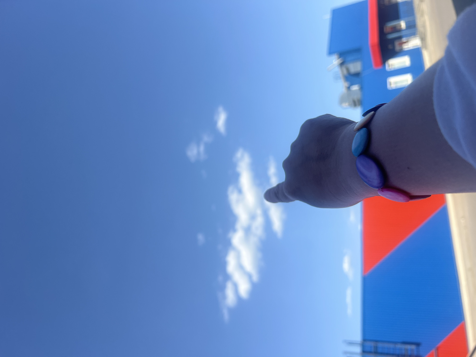 The image depicts a blue sky with scattered white clouds. A hand is pointing towards the clouds. The person is wearing a bracelet with multi-colored beads. In the background is a building with a red and blue facade, and white windows. The image was taken in the city of Chaikovsky, Russia.