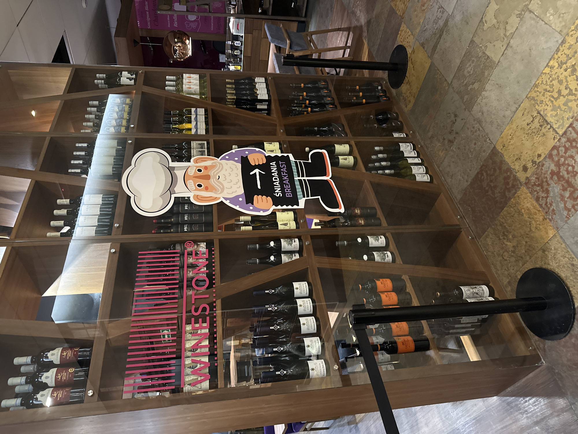 The image shows a display case filled with various wine bottles. The case is made of wood and has shelves for the wine bottles. On one of the shelves, a sign reads "ŚNIADANIA BREAKFAST". Another sign on the case reads "VINESTONE" and has the varietals “PINOT NOIR” “CANESTRARI” “CANISTRU”, “GRIFON”, “Chi”, “Sauvignon Blanc”, “Chardonte”, “LOC”, “Rieslin”, “Viognie”, “Pin” with a number “192” in front of the wines. The floor is tiled with a geometric pattern. The photo was probably taken in a shop in Wrocław, Poland that offers food and wine.