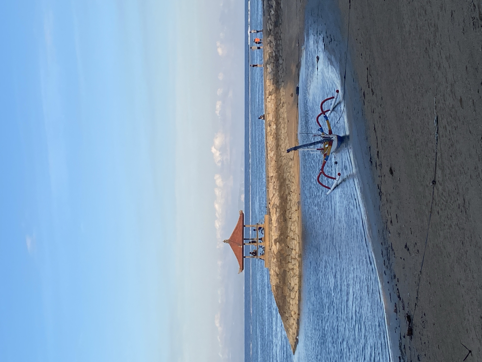 The image captures a serene coastal scene at Sanur Beach in Denpasar, Indonesia. The bright blue sky is adorned with scattered white clouds. The sea, a deep blue, meets a sandy beach where a traditional Balinese outrigger boat, painted in vibrant red and yellow, rests partially on the shore and partially in the water. In the middle ground, a man-made stone jetty extends into the sea, topped with a Balinese-style pavilion with a red, tiered roof. Several people are visible on the jetty, some appearing to be conversing or resting. The overall atmosphere is tranquil, suggesting a peaceful day at the beach.