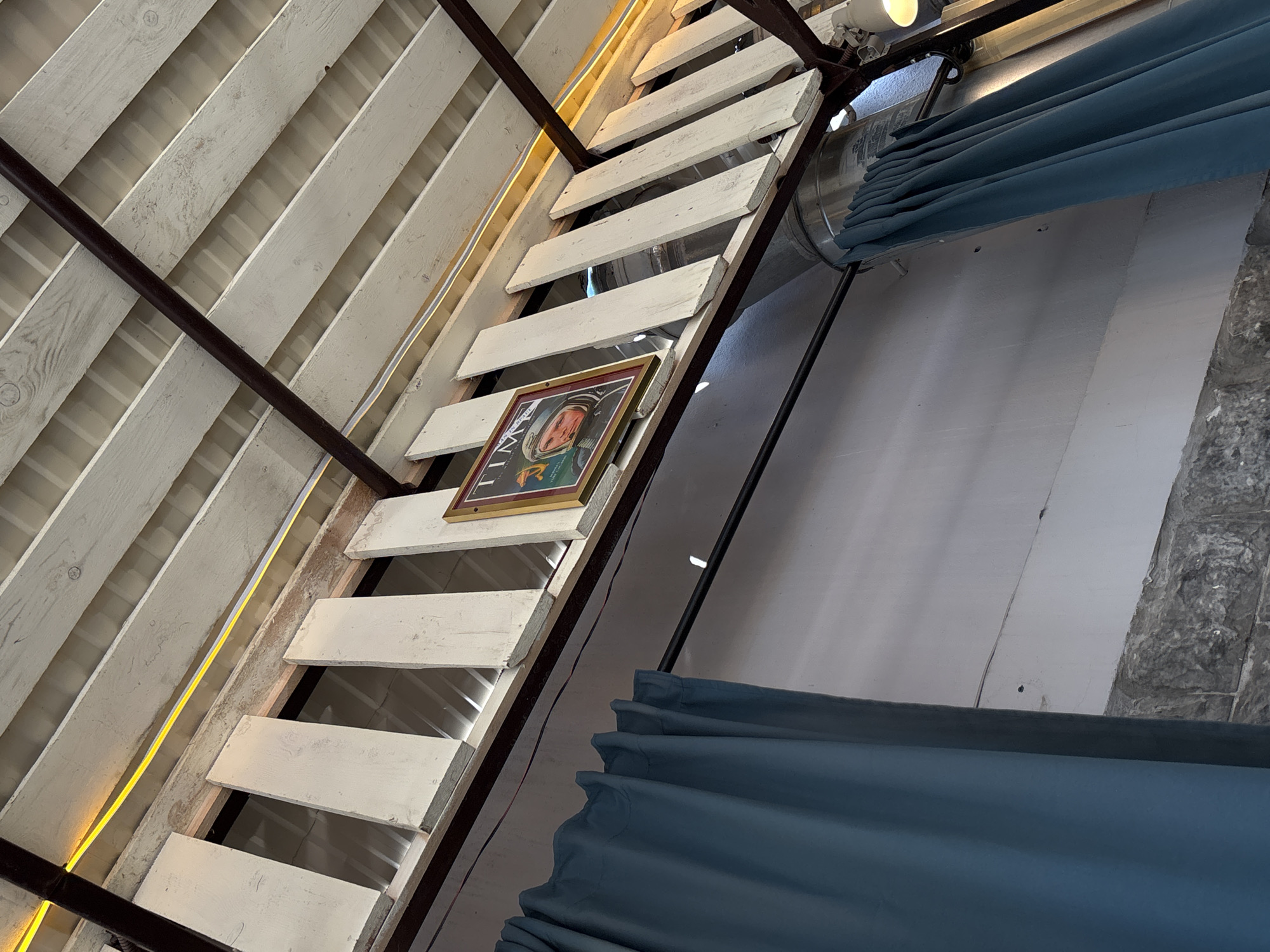 A low-angle shot shows a wooden staircase at an angle with white treads and brown support beams. A framed portrait hangs off to the side on the stairs. The environment includes exposed white wall paneling, stone or rock-like material, and hanging blue curtains. Light fixtures illuminate the scene. There is no visible indication of the location being Chaikovsky, Russia.