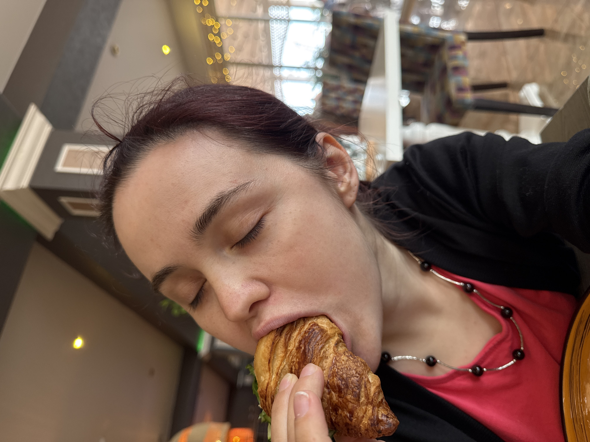 A woman is eating a croissant indoors. She has dark hair and closed eyes. She is wearing a black cardigan over a red shirt, and a beaded necklace. The background is blurry, but shows lights and architectural elements, suggesting an interior setting, potentially a cafe or restaurant. The city and country are not discernible from the image.