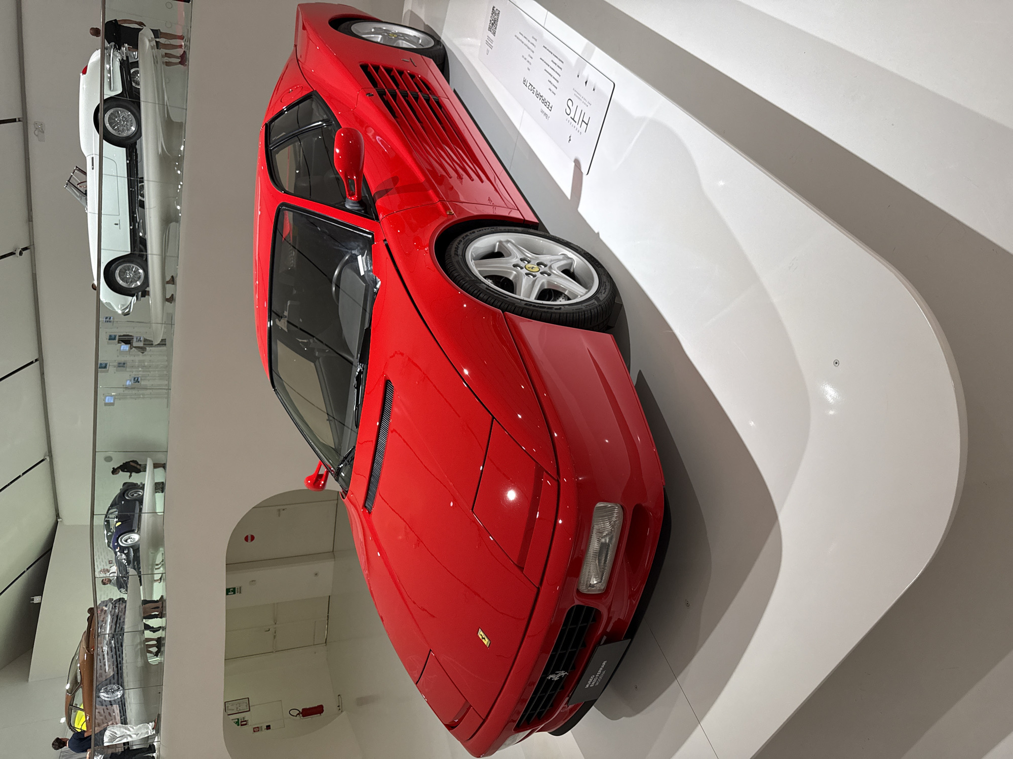 A vibrant red Ferrari sports car, specifically a Ferrari 512TR, is the central focus, displayed prominently on a curved white wall in a museum. The car is angled, with its front section closer to the viewer, showcasing its sleek lines, pop-up headlights, and a small yellow Ferrari badge. Distinctive horizontal vents are visible on its rear hood, and it sits on five-spoke silver wheels.
To the right of the red car, an information plaque is mounted on the white wall. Partially visible text on this plaque identifies the vehicle as a "FERRARI 512TR" and includes the word "HITS."
In the background, beyond a glass partition or reflective surface, other classic cars are displayed on what appears to be another level of the museum. A white convertible with spoked wheels is clearly visible, along with reflections of other vehicles, including a dark blue car and a brown car. Several visitors are also reflected in the glass, indicating an active museum environment.
The setting is a modern, well-lit indoor space, characterized by clean white walls and a minimalist aesthetic typical of an automotive museum. Given the location context of Modena, Italy, this is likely an exhibit celebrating Ferrari's heritage, such as the Enzo Ferrari Museum. The image captures a moment of automotive appreciation and history.