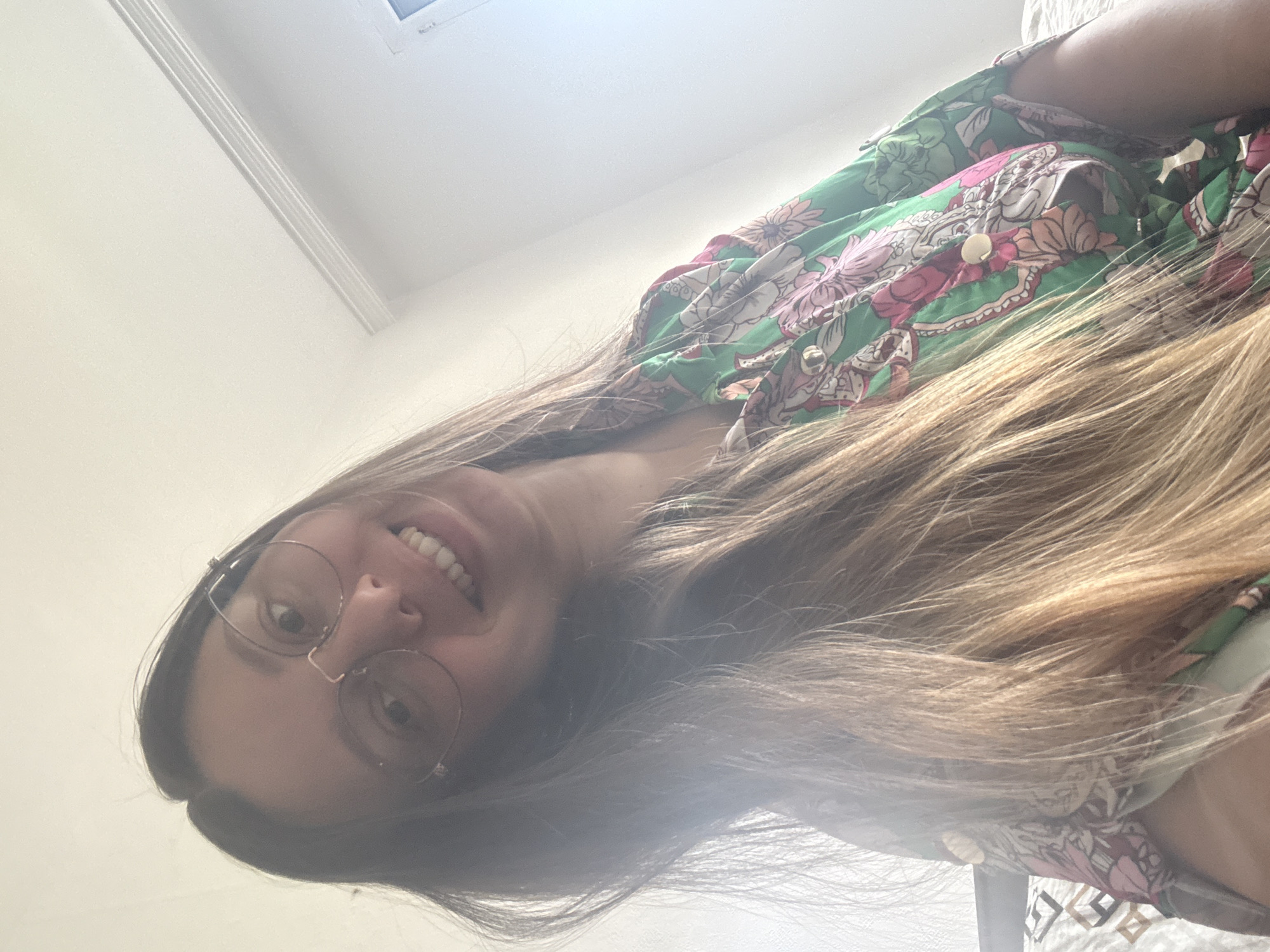This is a close-up, upward-angle selfie of a young woman with long, blonde-brown hair, wearing large, round eyeglasses and a green button-up shirt with a floral pattern. She is smiling and her head is tilted. The background is a plain, light-colored wall with some decorative molding near the ceiling. The image appears to be taken indoors, likely in a bedroom. There is no identifiable information about the location in Plovdiv, Bulgaria, or any other city.