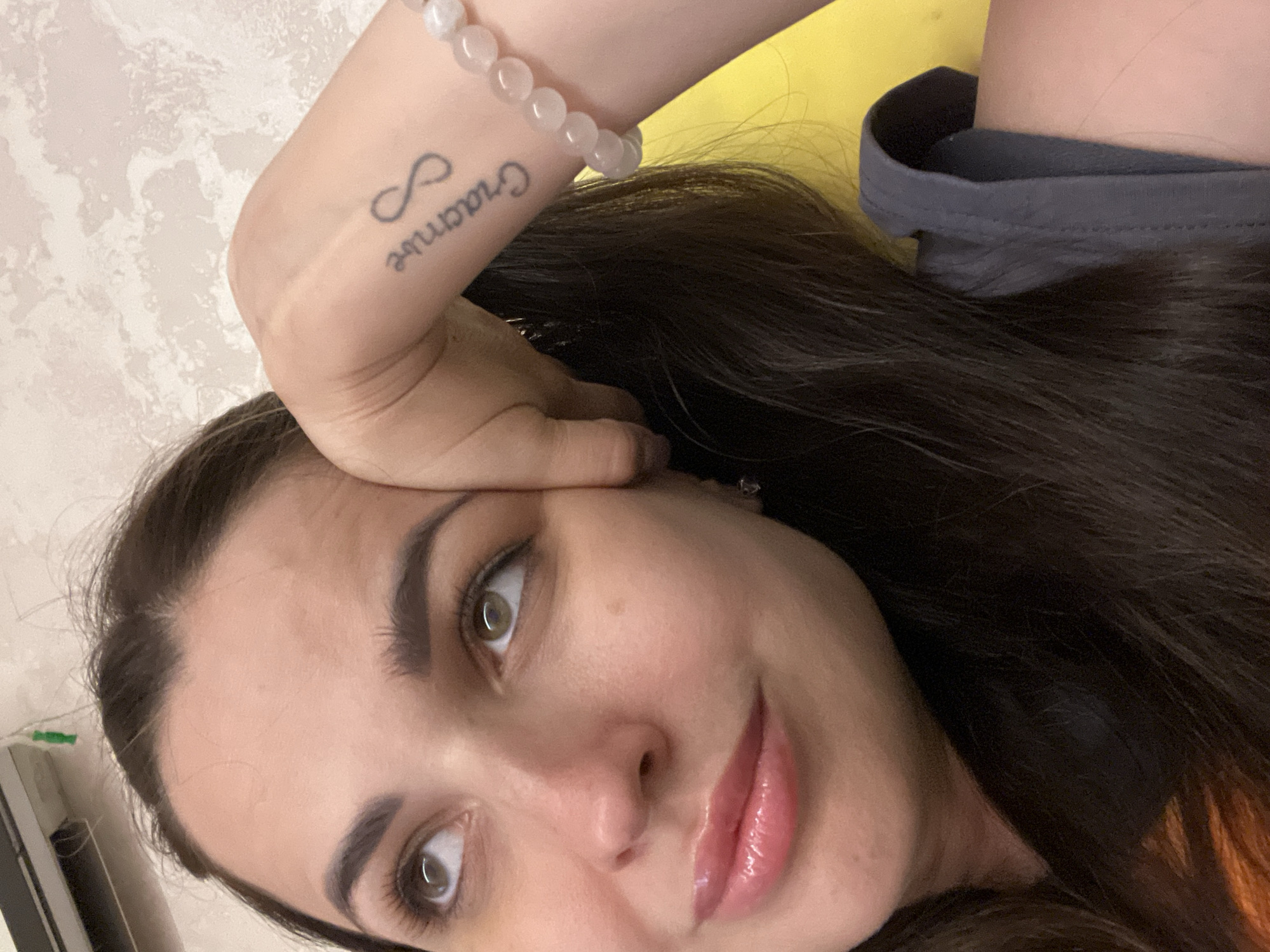 A close-up, medium shot shows a woman with dark hair looking upwards and to her left, resting her head on her left hand. Her eyes are wide and her lips are parted slightly, with a sheen of gloss on them. She is wearing a dark gray top. On her left forearm, a tattoo of an infinity symbol is visible above some cursive text. A light pink beaded bracelet is worn on her wrist. The background consists of a textured wall and a bright yellow object, possibly a cushion or a piece of furniture. The scene is lit by artificial lighting.