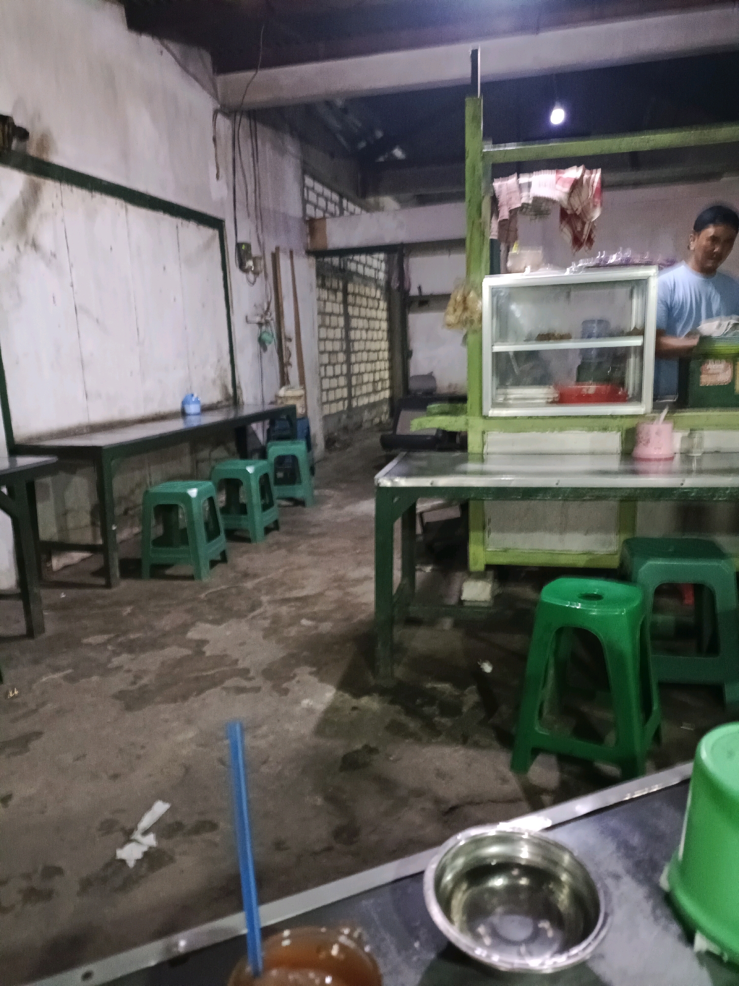 The image depicts an indoor food stall in Sukomulyo, Indonesia. A male individual, wearing a light blue t-shirt, is visible in the background behind a glass display case. The stall features green plastic stools and metal tables. The flooring appears to be concrete. In the foreground, a metal bowl and a blue straw are visible on a metal surface. The lighting suggests the stall is operational.