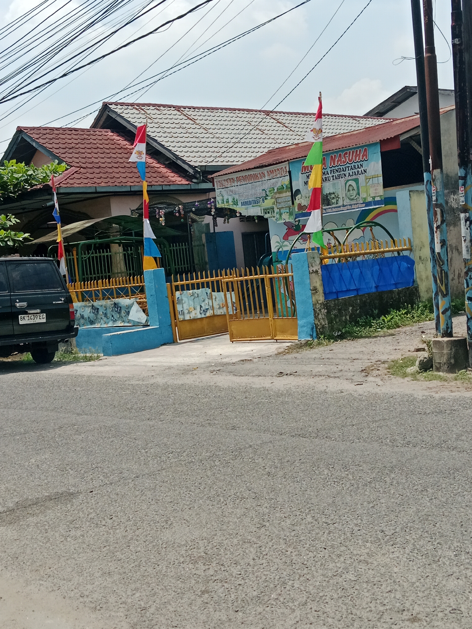 The image shows a building with a red tiled roof and a grey tiled roof, possibly a school or community center, located in the City of Binjai, Indonesia. In front of the building is a blue and yellow fence, and a yellow gate. There are several flags flying, including the Indonesian flag. A black SUV is parked on the left side of the image, with a license plate that reads "BK 1439 EG". A banner on the building reads "NASUHA" and displays information about school registration. The area appears to be a public street with power lines overhead.