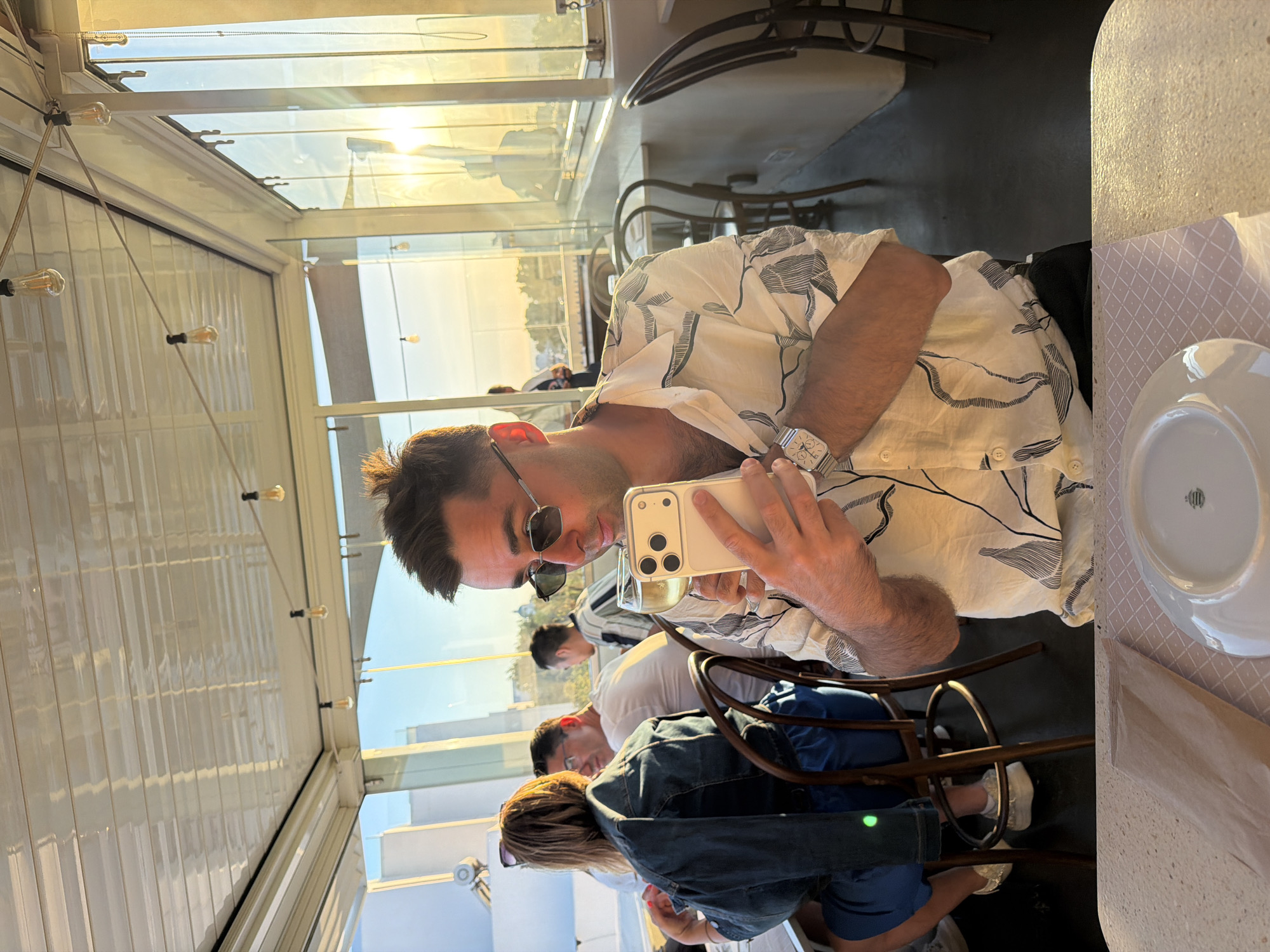 In an outdoor dining setting, bathed in the warm glow of sunset, a man in a white patterned shirt and sunglasses holds a white smartphone and a glass of white wine. He is seated at a table with a white plate and a brown paper bag. Behind him, a woman in a denim jacket sits at another table. The background reveals a glass-enclosed area with tables and chairs, and beyond that, a city landscape. The location is identified as a Municipal Unit in Ia, Greece.