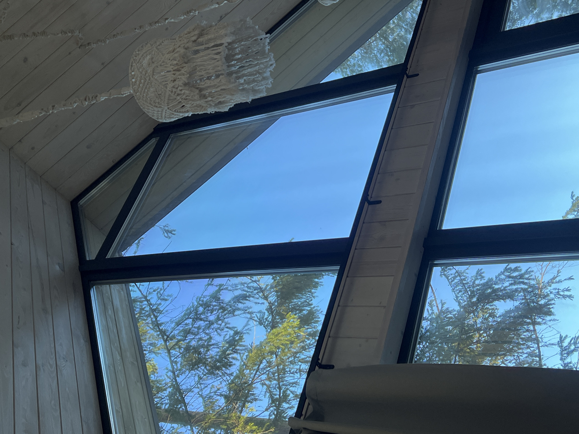 The image presents an interior view through a set of geometrically arranged windows. Outside, the sky is visible through some windows, indicating a clear day. Trees with green foliage are seen in the lower windows, suggesting a natural or suburban environment. The interior is characterized by light-colored wooden paneling on the walls and ceiling. A decorative object, possibly a light fixture or hanging ornament, is suspended from the ceiling. The window frames are dark and contrast with the lighter wood. The location specified is Мишкино, Russia, but this cannot be verified from the image alone. There are no people visible in the frame.