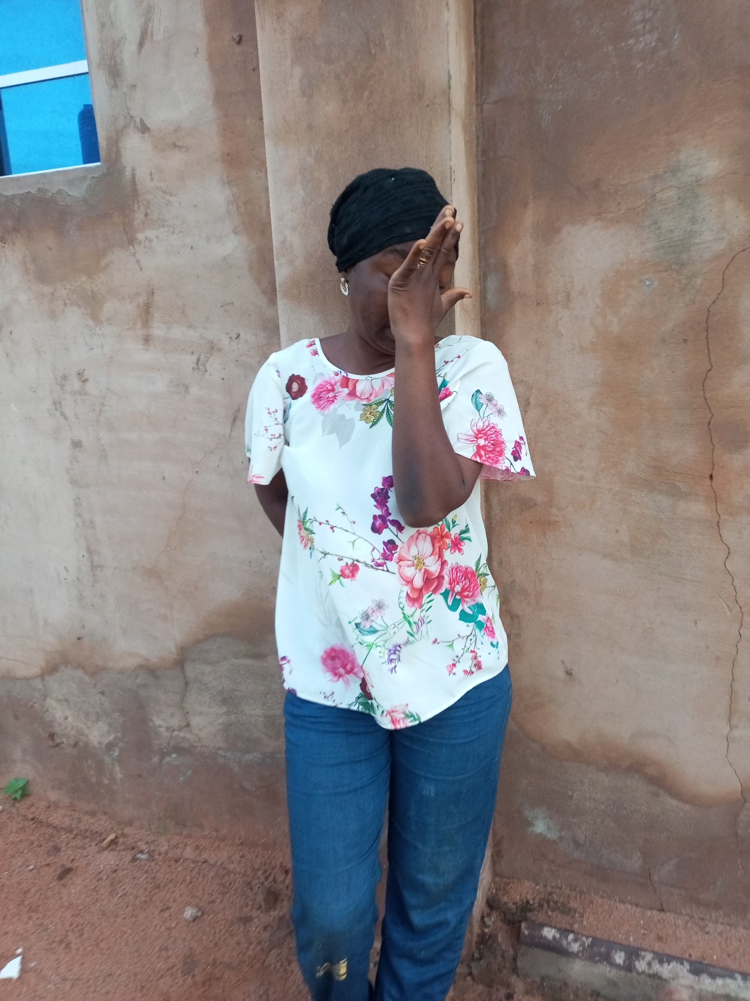 A full-shot, eye-level medium-wide photograph shows a dark-skinned woman with a black head covering standing in front of a textured, tan-colored wall. The wall has some visible cracks and discoloration, suggesting an outdoor environment. To the left of the wall, a section of a blue-framed window is visible. The woman is wearing a white short-sleeved blouse with a floral pattern of pink, purple, and green flowers. She is also wearing blue jeans. Her right hand is raised, partially covering her face as if shielding her eyes from the sun or in a gesture of distress. Her left arm is bent behind her back, with her hand out of view. The ground in front of her appears to be dirt or sand, with some small green foliage on the left. The lighting suggests it is daytime, possibly with bright sunlight casting shadows. There are no other people visible in the frame.