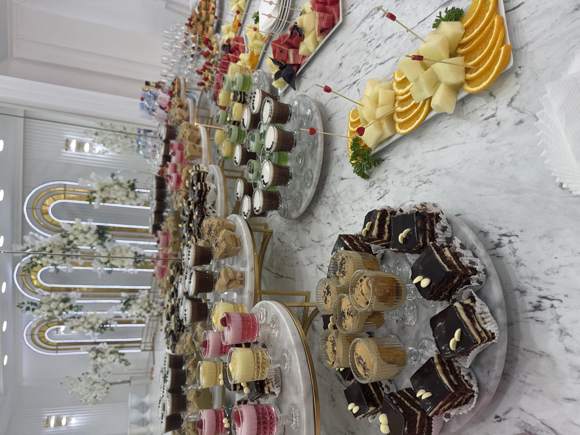 The image depicts a dessert and fruit buffet at a venue, likely for an event, in Mălăiești, Moldova. The table is laden with an array of sweets, including individual layered chocolate cakes, small glasses filled with layered desserts or mousses, and shot glasses containing colorful layered beverages or custards. There are also platters of fresh fruit, such as sliced melon and oranges, garnished with parsley and decorative picks. In the background, a decorative archway with floral arrangements and lighting suggests a festive or elegant setting. No people are visible in the frame. The overall environment appears to be indoors, with a marble-patterned tabletop and white walls.