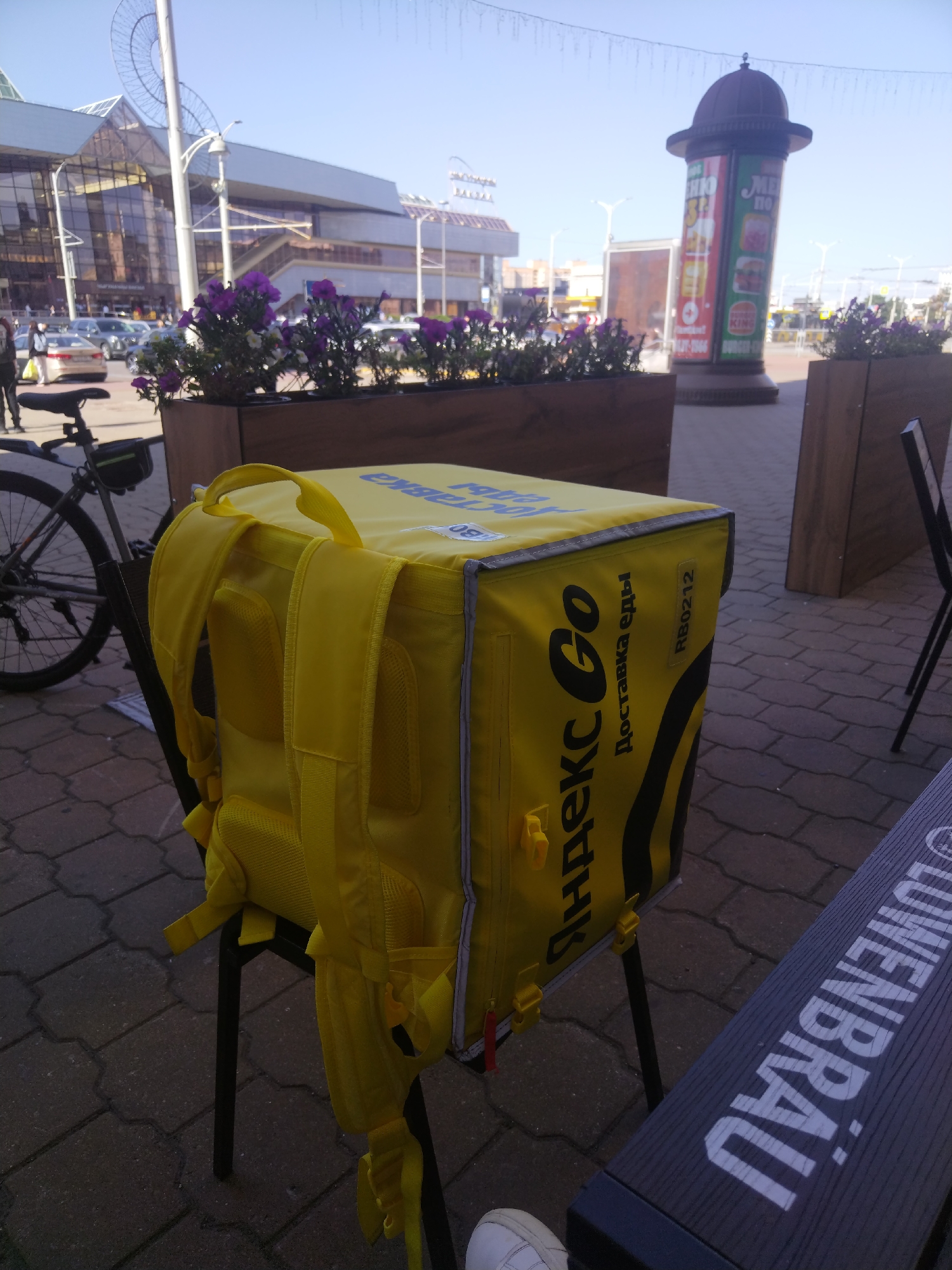 An exterior shot captures a bright, sunny day in Minsk, Belarus, with the distinctive architecture of the "Minsk-Arena" visible in the background. In the foreground, a yellow delivery backpack, emblazoned with the "Yandex Go" logo, sits on a black chair, suggesting a courier is nearby. A bicycle is parked to the left of the backpack, also indicating a delivery service. The scene is further enhanced by planters filled with purple flowers, adding a touch of nature to the urban landscape. A tall cylindrical billboard, likely advertising Burger King, stands prominently in the mid-ground. The ground is paved with interlocking stone tiles, typical of city sidewalks. A dark bench with white lettering, possibly advertising Löwenbräu, is situated in the lower right corner of the frame. The overall impression is one of a bustling city, with everyday life and commerce taking place under a clear blue sky.