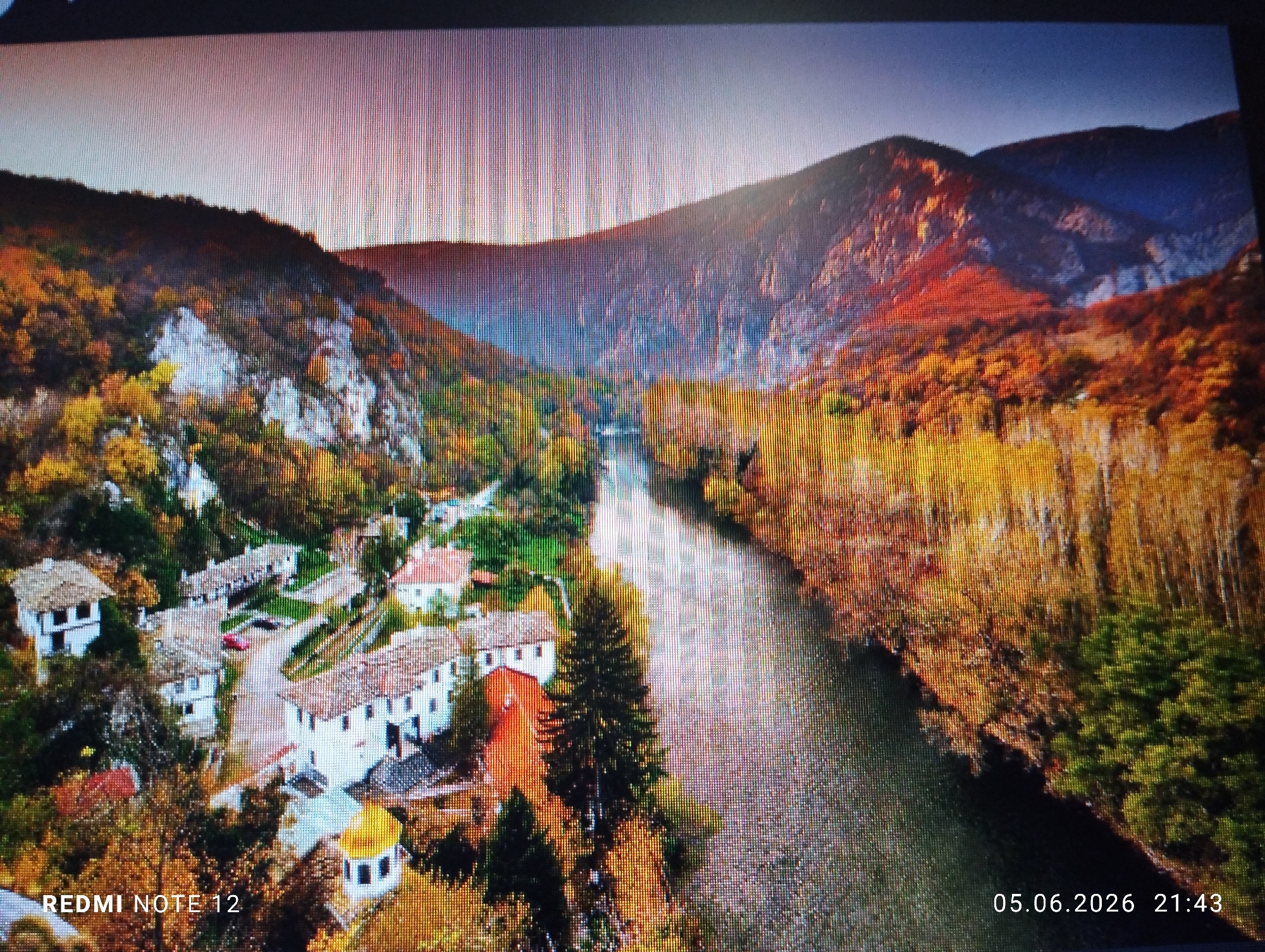 The image is a photograph of a digital screen displaying a panoramic aerial view of a mountainous river valley during autumn. The scene depicts a wide river flowing through the center of a valley, bordered by steep, forested mountains on both sides. The foliage on the trees displays vibrant autumnal colors, including shades of yellow, orange, and red, with some darker green coniferous trees also present. Exposed, light-colored rock faces are visible on the mountainsides, particularly on the left.
On the left bank of the river, a compact village or settlement is visible, characterized by multiple white-walled buildings with reddish-brown tiled roofs, indicative of traditional architecture. A distinctive small structure with a golden-yellow dome is located among the buildings. A single red vehicle is faintly discernible on a pathway within the village area. No individuals are visible within the scene.
The sky above the mountains is clear, transitioning from a lighter, almost white or pale pink hue near the horizon to a deeper blue at the top right.
The image itself contains digital watermarks indicating the device that captured the screen photo ("REDMI NOTE 12" in the bottom left) and a timestamp ("05.06.2026 21:43" in the bottom right).
Based on the visual evidence of a prominent mountainous landscape and river valley, this location does not correspond to the city of Burgas, Bulgaria, which is a coastal city. The scene depicts a mountainous region, likely within Bulgaria, but not Burgas itself.