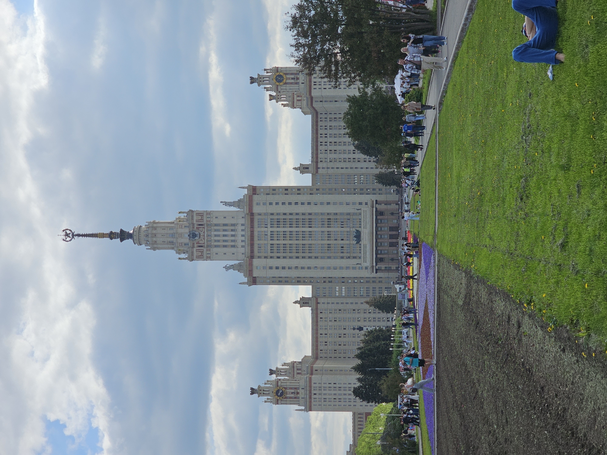 Here is a description of the image:
The image depicts the main building of Moscow State University in Moscow, Russia. The building is tall and imposing, with a distinctive spire topped by a star. It's framed by a partly cloudy sky. A grassy area and a flowerbed are in the foreground with people walking by. Someone is also relaxing on the grass.