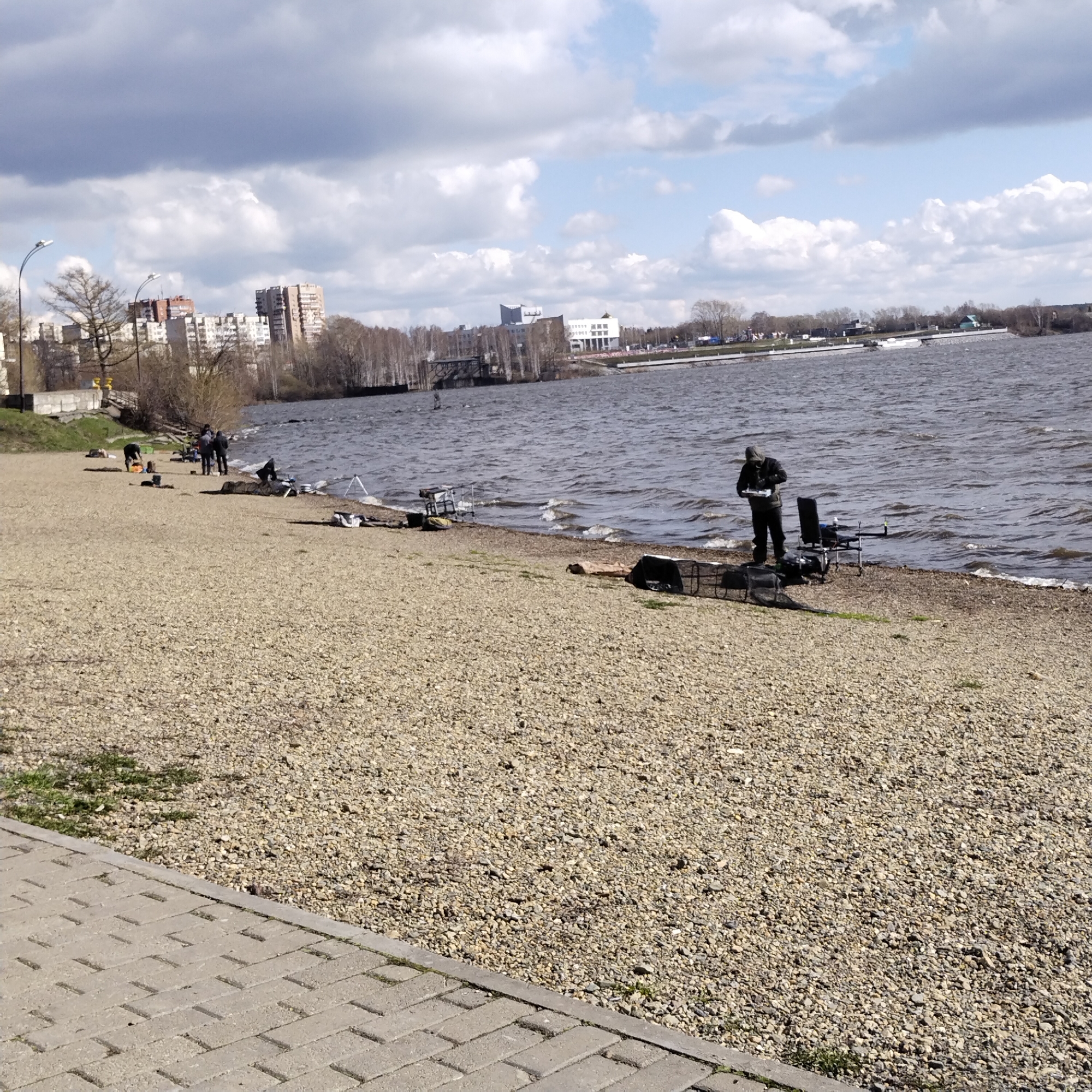The image depicts a pebbled beach along a body of water with several people fishing. The water is choppy, with small waves visible near the shore. In the background, buildings are visible, suggesting an urban setting. The sky is overcast with scattered clouds.
Specific details:
People: Several people are visible, some standing and fishing, others appear to be setting up or tending to their fishing equipment. The attire of the person closest to the camera suggests cool weather.
Subjects/Objects: Fishing rods, chairs, and other fishing gear are scattered along the beach. A concrete walkway is in the foreground.
Actions/Interactions: People are engaged in fishing activities.
Environment: The scene is an urban beach. The location is identified as being in the city of Nizhny Tagil, Russia based on visual similarities.