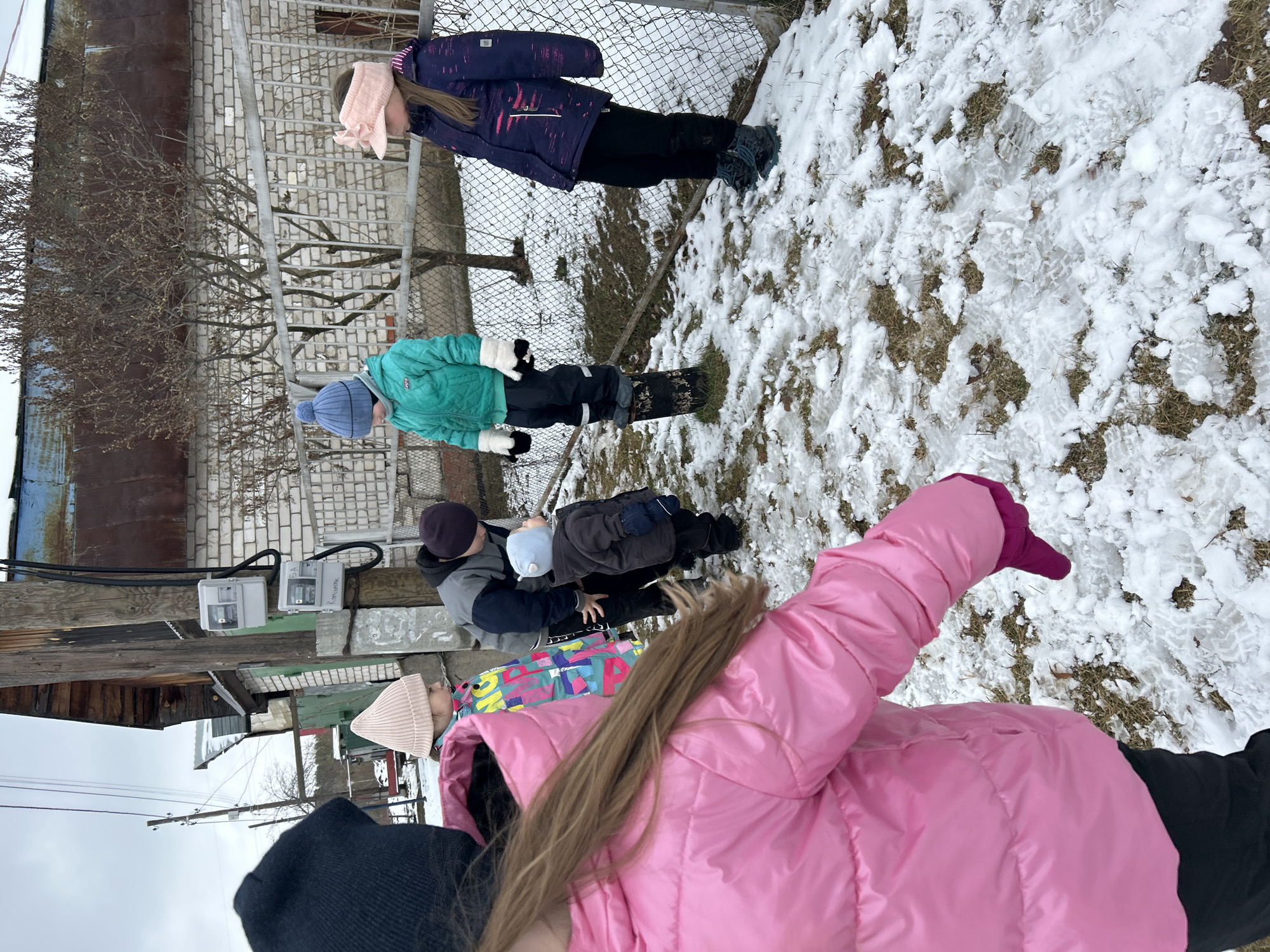 A group of five children of varying ages are standing outside in the snow near a chain-link fence, possibly in the city of Chaikovsky, Russia. The ground is covered in patches of snow and brown grass. The children are dressed in winter clothes, including jackets, hats, and mittens. From left to right: A child in a black hooded jacket and tan beanie looks into the scene; a child in a pink jacket with long blonde hair is facing forward; a child in a dark jacket and light blue hat is next to an adult; a child in a green jacket and blue hat stands further away; and a child in a purple jacket and pink headband is closest to the fence. The children are interacting with each other. Behind them is a brick building with electrical meters and bare trees.