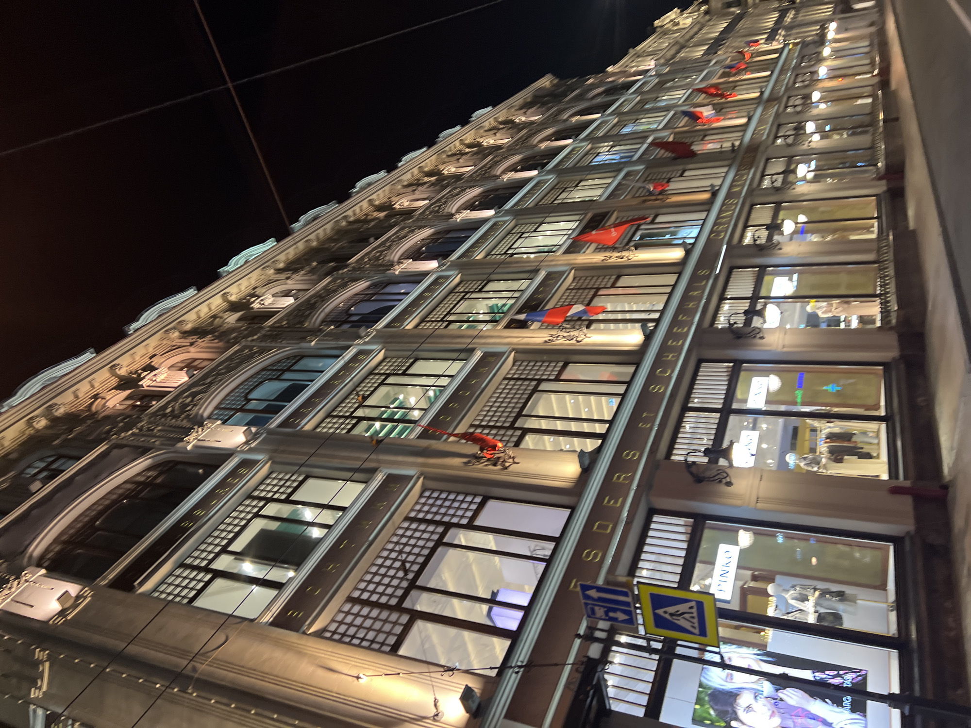 A low-angle, night photograph shows a tall building, identified as the "Traders and Scheerers" building, located in Saint Petersburg, Russia. The building, illuminated by lights, features a series of windows, some of which display mannequins and products. Red and blue banners adorn the facade between windows. A street sign, with arrows and a pedestrian crossing symbol, stands in the foreground. Overhead, electrical wires are visible against the dark sky.