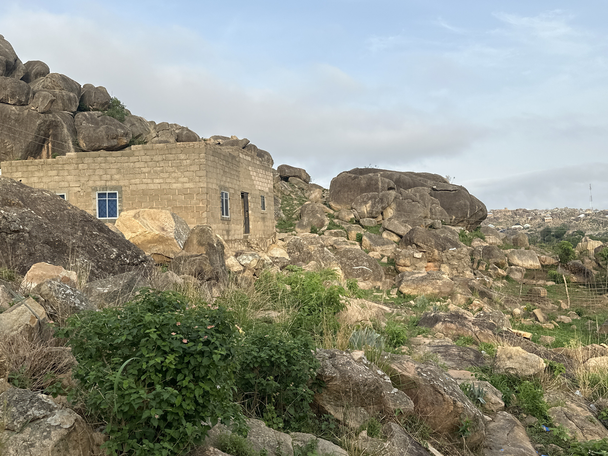 The image depicts a rocky landscape with a partially constructed building in Jos, Nigeria. The building is made of cinder blocks and has two visible window openings with blue-tinted glass and a doorway. The building is nestled among large, grey boulders and sparse vegetation consisting of dry grass, green shrubs, and succulents. The sky is overcast with grey and white clouds. In the background, a hillside is dotted with buildings and a tall antenna is visible on the right. The foreground is dominated by the rocky terrain and vegetation.