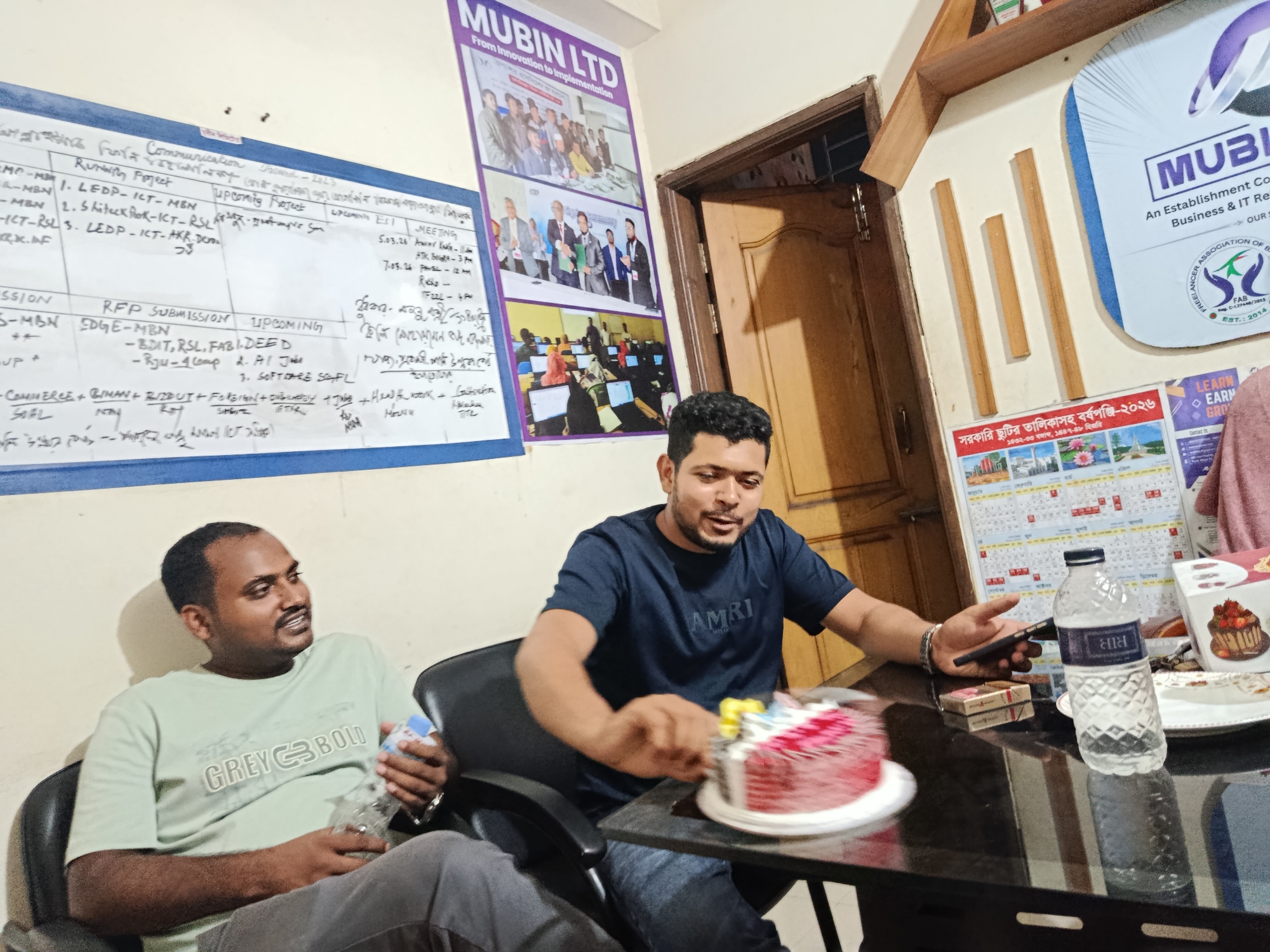 **Scene:** An indoor office setting in Dhaka, Bangladesh, with two men present.
**People:**
* Two men are in the scene. One sits in the left part of the frame wearing a light-green t-shirt with "GREY BOLD" printed on it. He is holding a water bottle. The second man wears a dark-blue t-shirt with "AMRI" printed on it. He is holding a smartphone and appears to be slicing a cake.
**Objects:**
* A cake is on the table.
* A water bottle is on the table next to a pack of cigarettes.
* A calendar is visible on the wall.
* A whiteboard with notes and schedules is on the wall.
* A poster/signage for "MUBIN LTD" is present on the wall.
* A promotional sign for "Free Lancer Association of Bangladesh" is also visible.
* A bottle of water is on the table.
**Actions:**
* The man in the dark-blue t-shirt is likely cutting a cake.
* The man in the light-green t-shirt is sitting and holding a water bottle.
**Interactions:**
* There is an interaction between the two men around the table
**Environment:**
* The scene appears to be in an office or work environment.
* The room has posters and schedules on the wall, likely related to work or business operations.