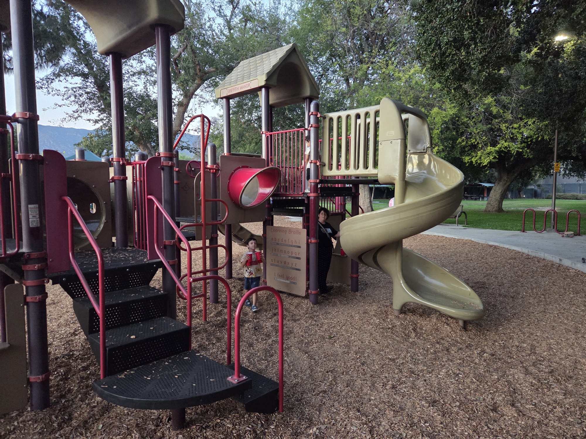 The image shows a playground structure at Griffith Park, Glendale, California. The structure is made of plastic and metal, with slides, stairs, and climbing elements. Two children are present, one a young boy standing near a sign, and another person, possibly an adult, standing to the right of the boy. The ground is covered with wood chips. In the background, trees and mountains are visible.
