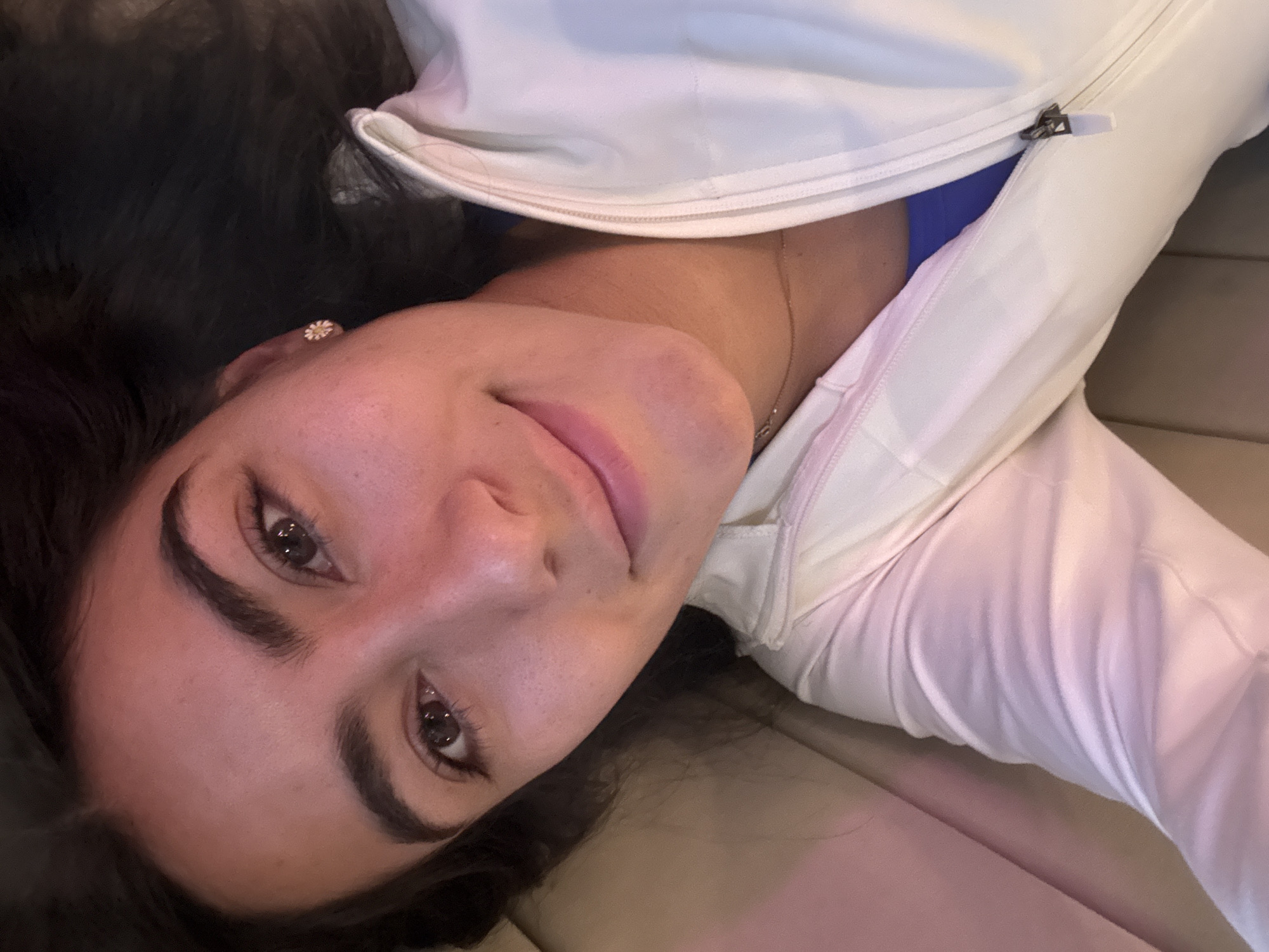 A young woman with dark, flowing hair and expressive dark eyes is depicted lying down, looking directly into the camera with a gentle, soft smile. Her head is tilted, giving the impression of a relaxed, informal pose. She has pronounced, well-groomed dark eyebrows.
She is wearing a light-colored, possibly white or very light pink, zipped jacket or hoodie. A hint of a blue garment is visible underneath the jacket's zipper. A delicate gold-toned necklace adorns her neck. In her right ear, a small, distinctive stud earring shaped like a white and yellow daisy or flower is visible.
The background consists of light brown or beige upholstery, suggesting she is reclining on a couch, bench, or similar soft furnishing. The lighting is soft and even, without harsh shadows, indicating an indoor setting, possibly during the day or evening with artificial illumination.
The scene captures a calm and engaging moment, likely a casual portrait or selfie taken in Puebla, Mexico. There is no visible text in the image.