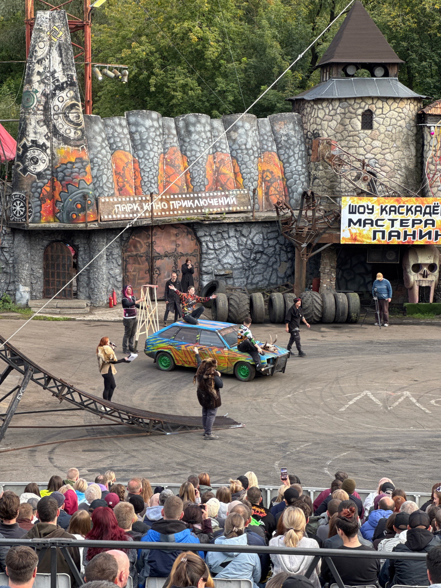 In Moscow, Russia, at the Park of Cinema Adventures, a stunt show is in progress. The background features a themed stage design resembling a rugged stone castle with a tall, conical tower. In front of this structure, a brightly painted car, decorated with graffiti and animal horns on its hood, is positioned on a paved area. Several individuals are actively participating in the performance. One person is balanced on top of the car, while others are standing or moving around it. A ramp is visible to the left, suggesting it's part of the stunt setup. A crowd of spectators is seated in a bleacher-style seating area in the foreground, observing the event. The overall scene depicts an outdoor entertainment performance with a dramatic and action-oriented theme.