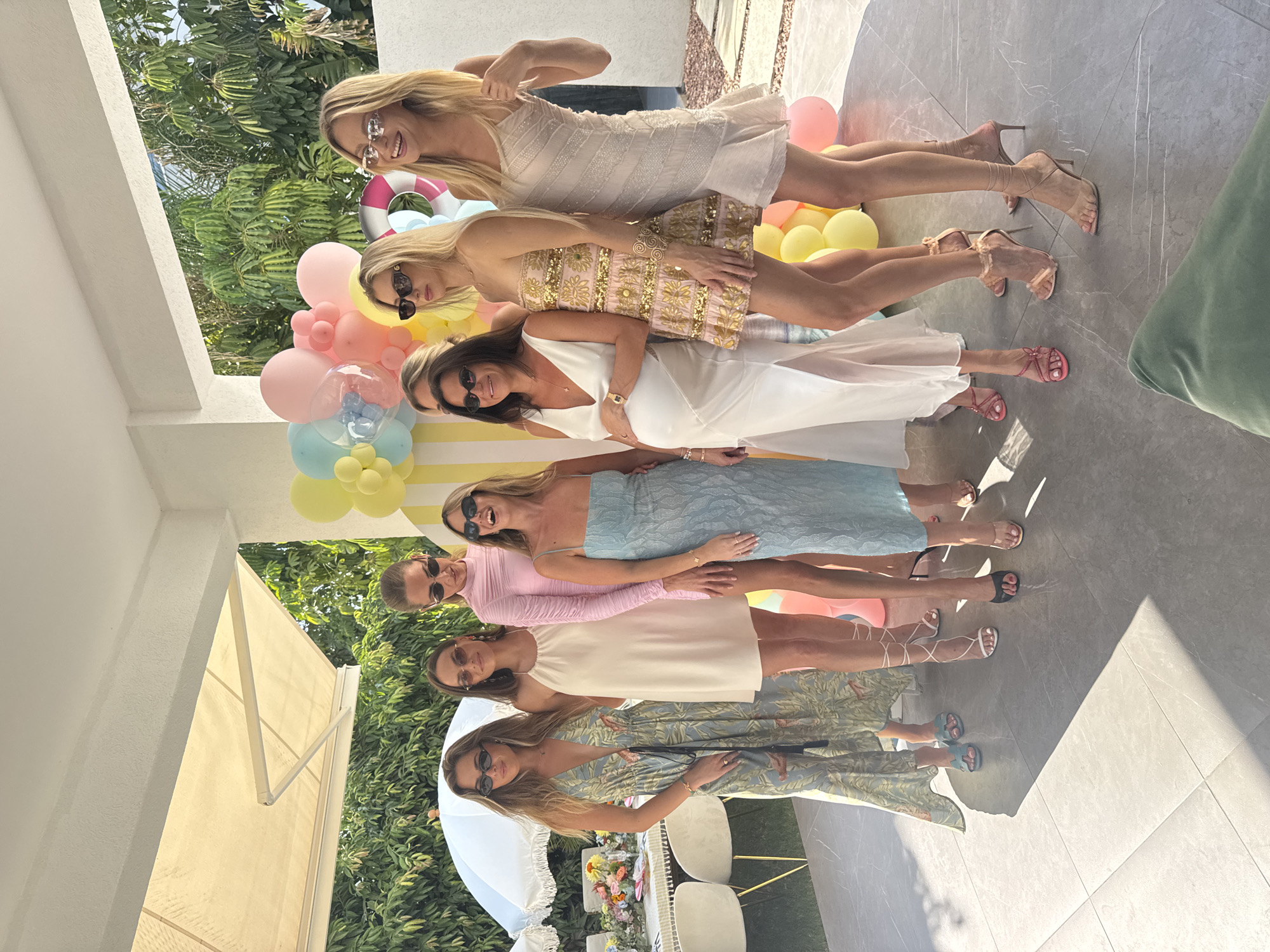 Eight women are posing for a photo outdoors in front of a balloon arch and a decorative display. The women are dressed in various fashionable outfits, including dresses and skirts, and are wearing sunglasses. They appear to be at a party or celebration. The location is a patio area with a tiled floor. In the background, there is greenery and trees, suggesting an outdoor setting. One woman is visibly pregnant. There is a table with floral arrangements and chairs visible to the left of the frame, indicating a dining or gathering space. The overall atmosphere seems festive and summery.