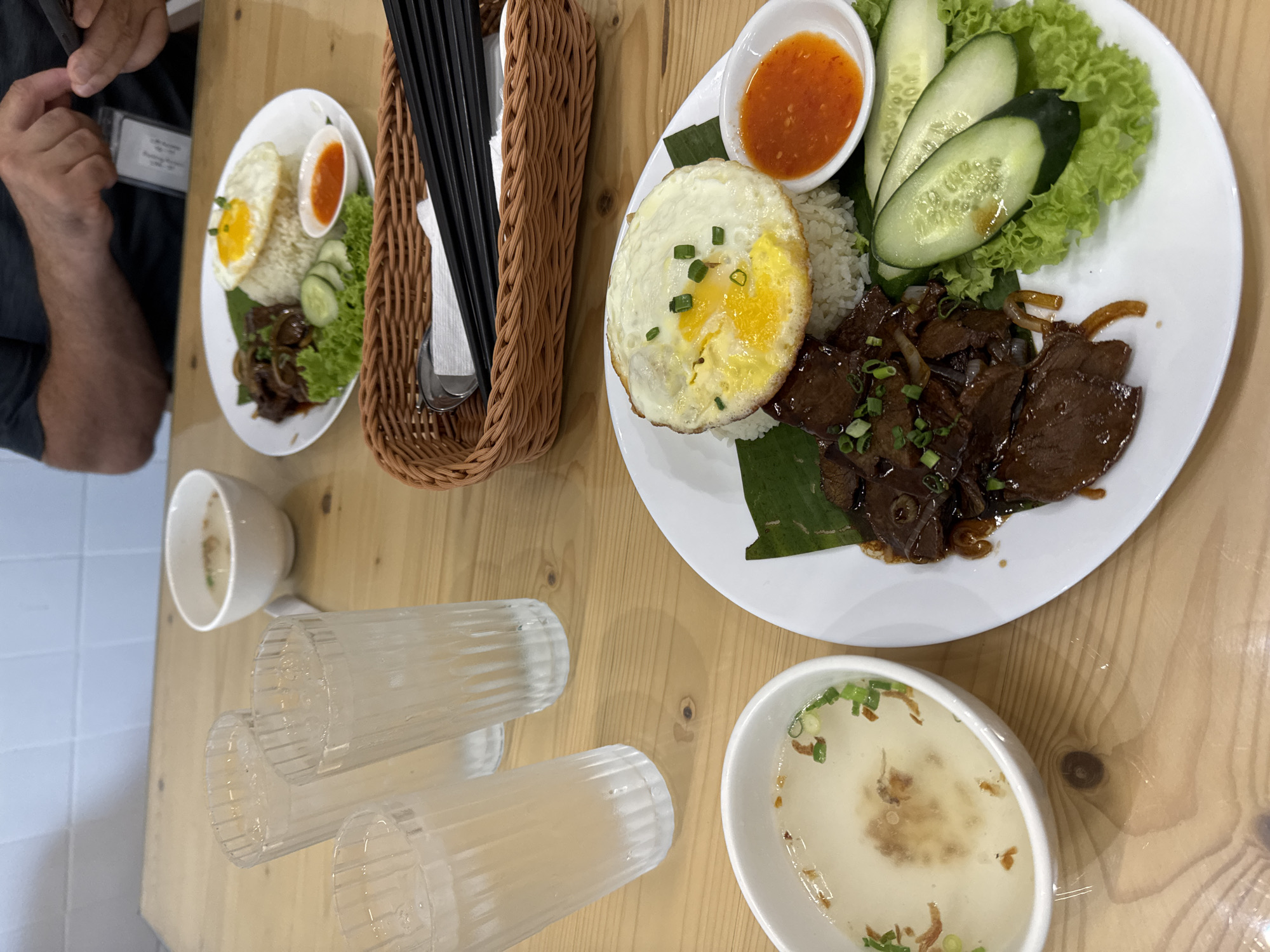 The image depicts a close-up, overhead view of a wooden table set with several dishes of food and beverages. In the foreground, a white plate holds a meal consisting of rice topped with a fried egg, a portion of dark meat, sliced cucumbers, and lettuce. A small dish of red sauce is also present on the plate. To the left, a white bowl contains a light-colored soup garnished with chopped green onions and fried shallots. Three tall, clear glasses filled with ice and a clear liquid, likely water, are arranged behind the food. In the background, another white bowl of soup and a plate with a similar meal are visible. A woven basket containing chopsticks and napkins is placed on the table. A person's hands are visible in the upper right corner, holding a smartphone. The overall setting suggests a casual dining environment, possibly a local eatery in Kuala Lumpur, Malaysia, given the common cuisine. The lighting is natural, coming from above, and highlights the textures and colors of the food.