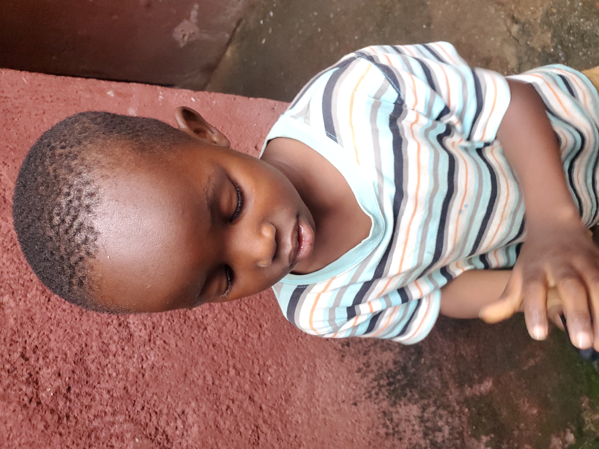 The image depicts a young Black child, likely male, with short dark hair and dark skin, looking downwards. The child is wearing a striped t-shirt with alternating bands of light blue, white, dark grey, and thin orange lines. The background consists of a textured, reddish-brown wall on the left and a lighter, rougher surface on the right, possibly another wall or building exterior. The ground in the lower right appears to be wet and possibly paved. The lighting suggests an outdoor setting, potentially during the daytime. The context of Jos, Nigeria, is provided, implying the scene is situated within this specific city. The child's hands are visible in the foreground, positioned close together as if holding or manipulating something small.