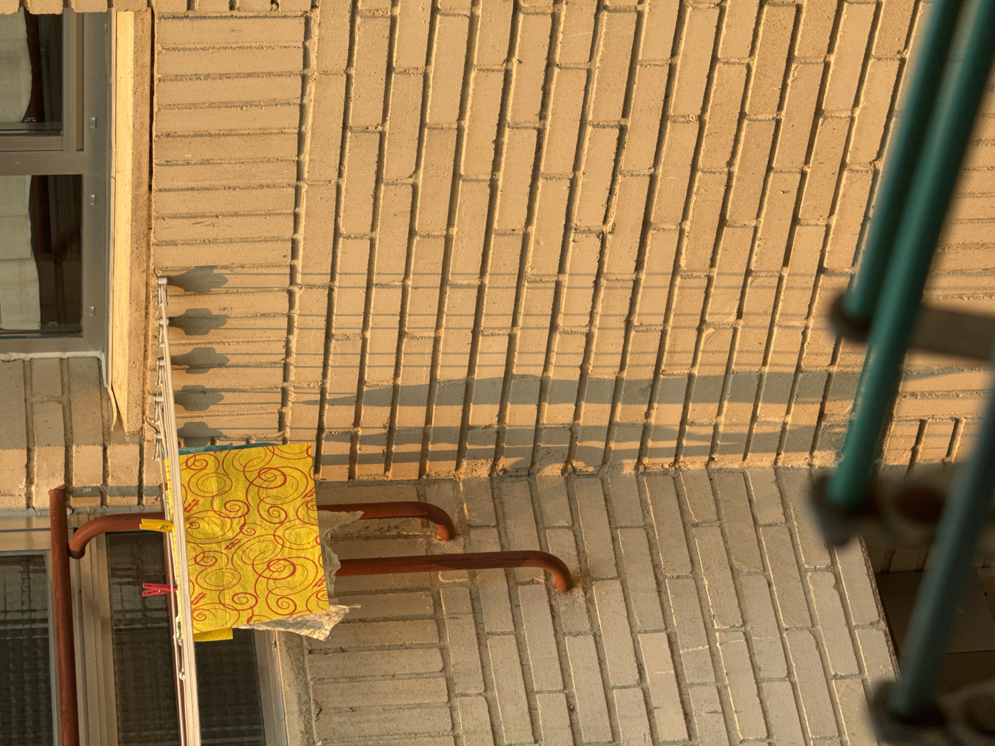 The image shows a close-up view of a brick wall with a clothesline strung across it. A yellow cloth with red spiral patterns is hanging from the clothesline, secured by pink and yellow clothespins. Two curved, reddish-brown metal pipes are attached to the wall below the clothesline. The wall is made of light-colored bricks and is casting a shadow from an unseen light source. In the foreground, a blurred green metal railing is visible. The scene appears to be an exterior wall, possibly on a balcony or a side of a building. The lighting suggests it is either morning or late afternoon, casting long shadows. The overall environment suggests an urban setting, consistent with Las Rozas de Madrid, Spain, a residential area. There are no people visible in the image.