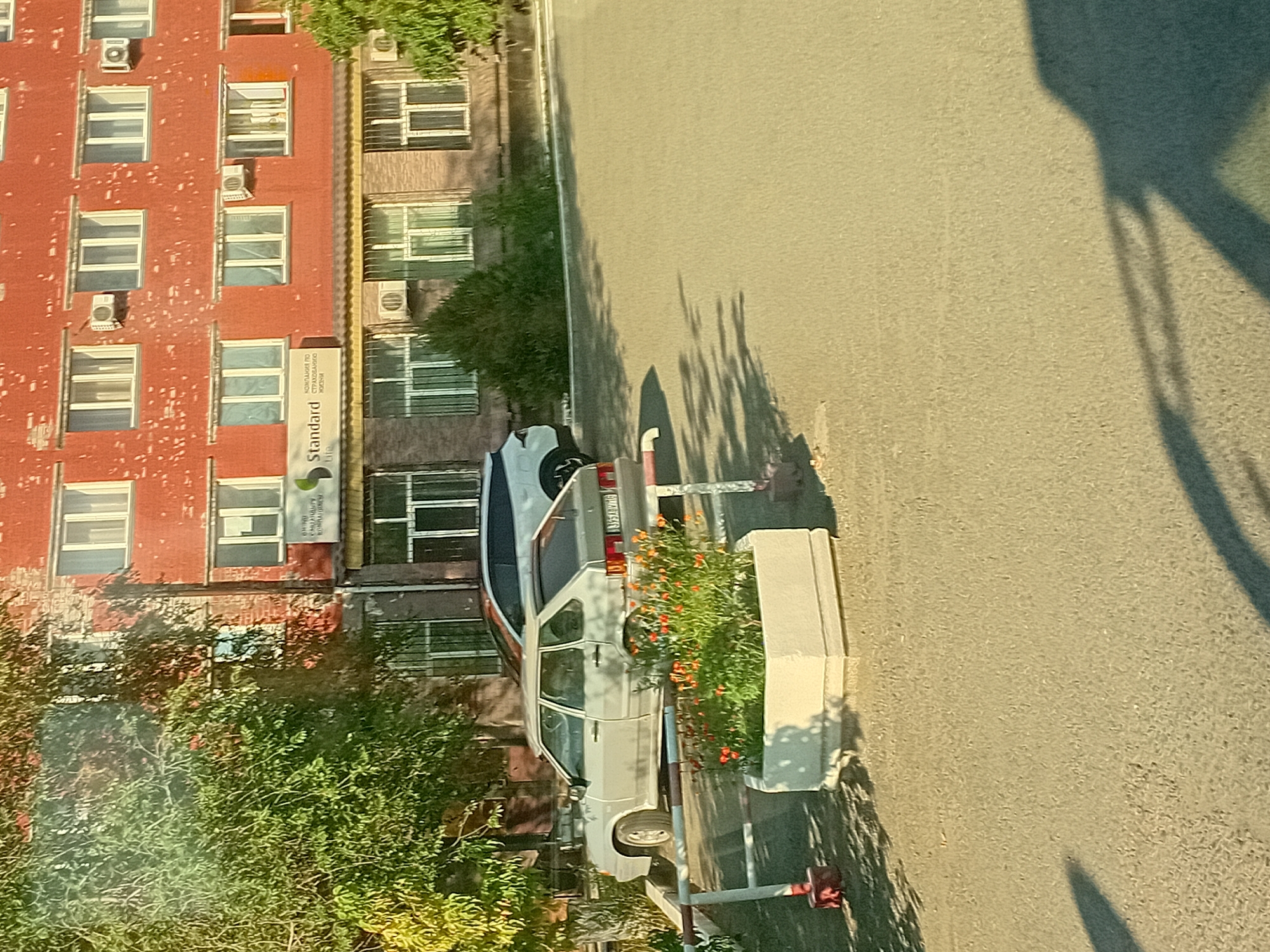 An outdoor, eye-level, medium shot captures a light-colored, five-door hatchback parked on an asphalt surface in front of a red brick building. The building has multiple windows and air conditioning units. A sign with green lettering and "Standard" in English is visible on the building. To the left of the parked car, a white planter with red flowers sits behind a white and red bollard. A shadow of a person is cast across the asphalt in the foreground. The image appears to be taken in the city of Semey, Kazakhstan, likely in a residential or commercial area.