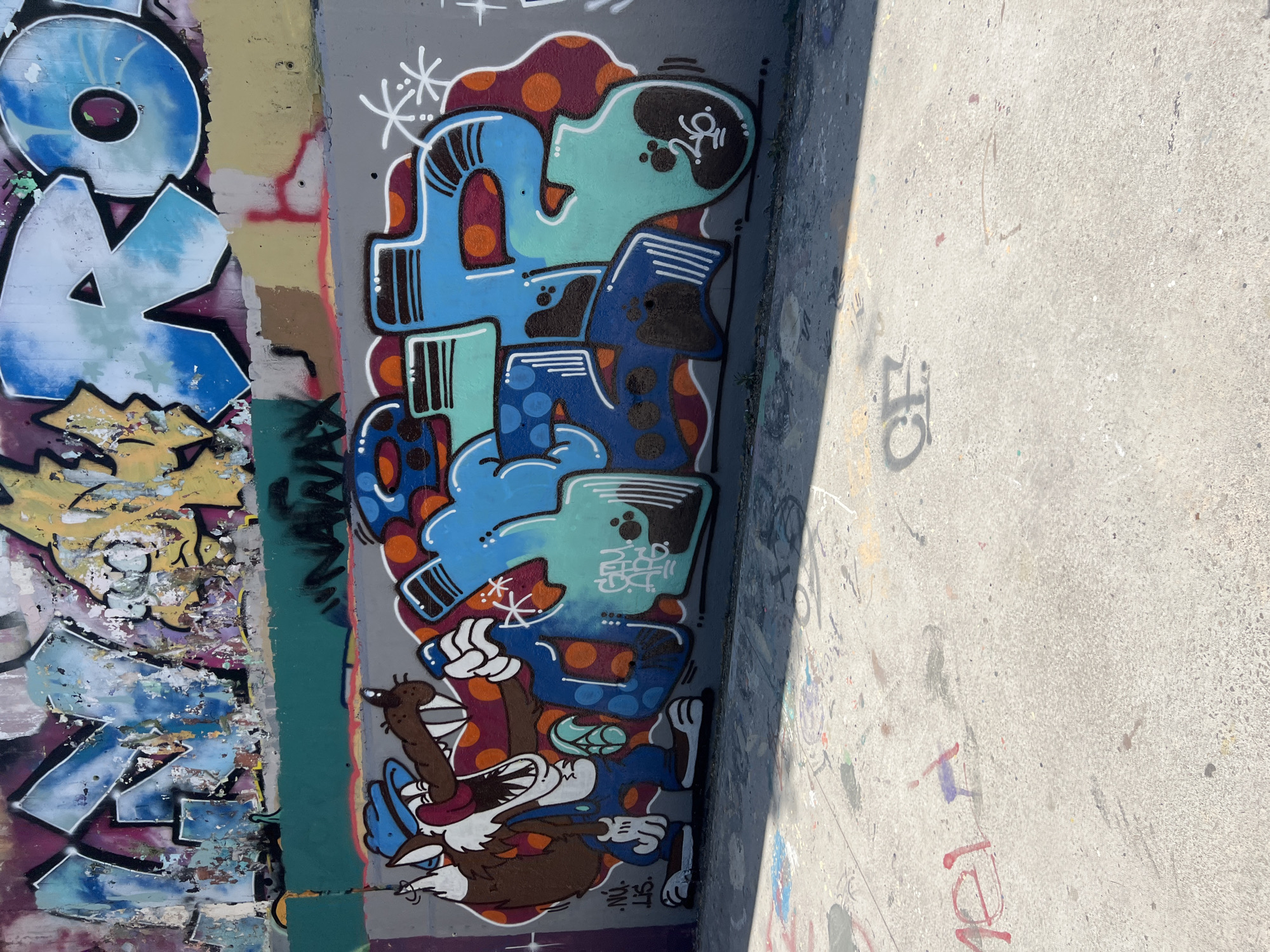 This media file captures a segment of graffiti art on a concrete wall and the adjacent ground. The graffiti on the wall is vibrant and colorful, featuring stylized cartoon characters and abstract lettering. On the left, large blue letters with white outlines, possibly part of a larger tag, are visible, with sections showing peeling paint underneath. To the right of this, a more intricate piece dominates, with a teal-colored head with black eyes and an open mouth, resting on a blue, segmented body. This figure is adorned with orange polka dots and dark blue sections with white stripes. A brown cartoon character, resembling a dog or fox with white gloves, is depicted in a dynamic pose at the bottom of this central graffiti. The graffiti on the ground is more subdued, consisting of faded tags and spray paint splatters in various colors.
The scene appears to be outdoors, likely in an urban environment, given the presence of graffiti and the concrete surfaces. The strong shadow cast across the scene, originating from the right and extending towards the left, suggests it is a sunny day, with the sun positioned high in the sky, possibly mid-morning or afternoon. The lighting indicates fair weather conditions. No specific events are depicted, but the graffiti itself suggests an artistic or subcultural expression. The overall impression is one of an urban art space, possibly a skate park or a designated graffiti wall, in Barcelona, Spain, as indicated by the location context. Visible text includes stylized letters within the graffiti art and faded tags on the concrete ground.