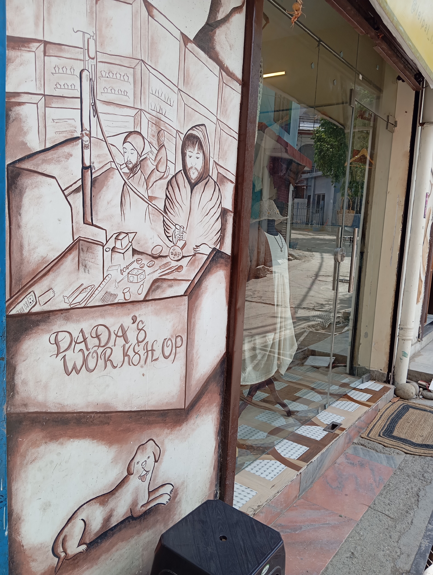 A street scene in Jonk, India, shows a shopfront with a mural on the left wall depicting "DADA'S WORKSHOP." The mural is brown and white and features two figures working at a table laden with tools and objects. Shelves are behind them, filled with more items. Below the table is a drawing of a reclining dog. The shop itself has a glass door and window, reflecting the street outside. A mannequin dressed in a white outfit is displayed inside the shop. A black plastic stool sits near the mural, and a doormat is to the right of the shop entrance. The street outside appears residential.