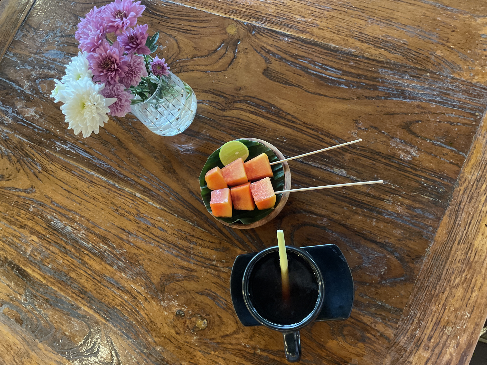 The image captures a scene on a weathered wooden table in the Ubud District, Indonesia. A small glass vase holds an arrangement of white and purple chrysanthemums. Next to it, a wooden bowl contains cubed papaya and a wedge of lime, skewered with wooden sticks. Below the papaya, a black mug filled with a dark liquid, possibly coffee or tea, is present. The mug sits on a black saucer, with a straw-like stick. The table's surface shows an intricate grain pattern, suggesting age and use. The overall composition suggests a casual, perhaps outdoor, setting with local food and drink.
