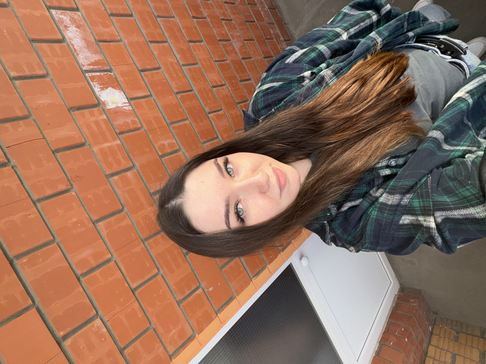 A young woman with long, dark brown hair and light blue eyes is taking a selfie in front of a red brick wall. She is wearing a dark green and blue plaid shirt over a grey t-shirt and light-wash jeans. A white door with a black screened window is visible to her left. The scene appears to be outdoors, with a concrete ground and a portion of a grey wall visible in the background. The lighting suggests it may be daytime, possibly overcast. The location is identified as Perm, Russia.