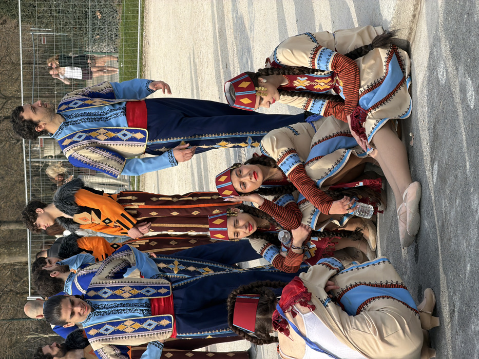 The image depicts a group of individuals in traditional folk costumes gathered in a park-like setting on a clear day in an unknown city. In the foreground, four females are seated or crouching on a paved surface. They wear long, light-colored dresses with patterned sleeves and blue panels, topped with red, flat hats adorned with yellow geometric designs and, for some, gold coin-like embellishments. Their hair is styled in long, dark braids. Two of the seated females hold clear plastic water bottles. One female is seen from the back, revealing a white tank top beneath her costume.
Behind the seated females, approximately five to six males stand, also in traditional attire. Their costumes consist of light blue long-sleeved shirts, dark blue trousers with vertical embroidery, and short, open-front vests featuring intricate blue, gold, and cream geometric patterns. They wear red sashes around their waists. One male in the mid-ground wears an orange jacket with black trim and a fur collar over patterned brown trousers.
In the background, a metal chain-link fence separates the paved area from a grassy space with trees. Several individuals in casual modern clothing are visible behind the fence, some observing the group. The lighting suggests a bright, sunny outdoor environment.
