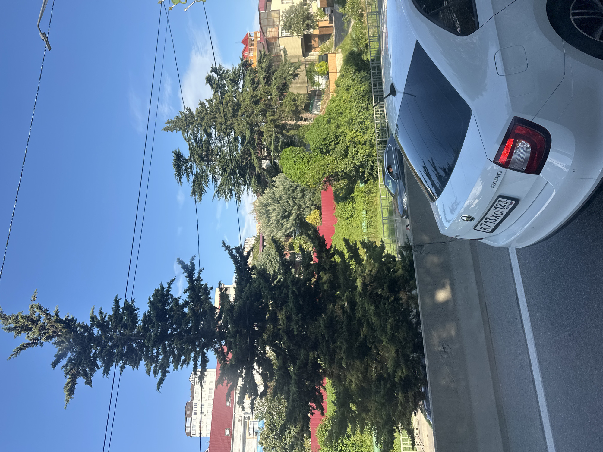 This is a medium-angle, outdoor, eye-level shot taken on a sunny day in Sochi, Russia. The foreground is dominated by the rear of a white Skoda Rapid sedan, its license plate "K713XO 123 RUS" visible. The car is on a road, separated from the background by a concrete barrier.
Behind the barrier, a lush green landscape with various trees and bushes thrives. Two large evergreen trees, possibly cypresses, stand prominently. A red fence runs horizontally across the middle ground, behind which are several buildings. One building, taller and lighter in color, has a red roof. Other buildings are visible further back, in shades of beige and yellow, with some featuring red roofs as well. Power lines are strung across the bright blue sky. In the background, another car, a dark sedan, is seen on a road, with a person in the driver's seat. The overall impression is a sunny day in a developed area with significant natural greenery.