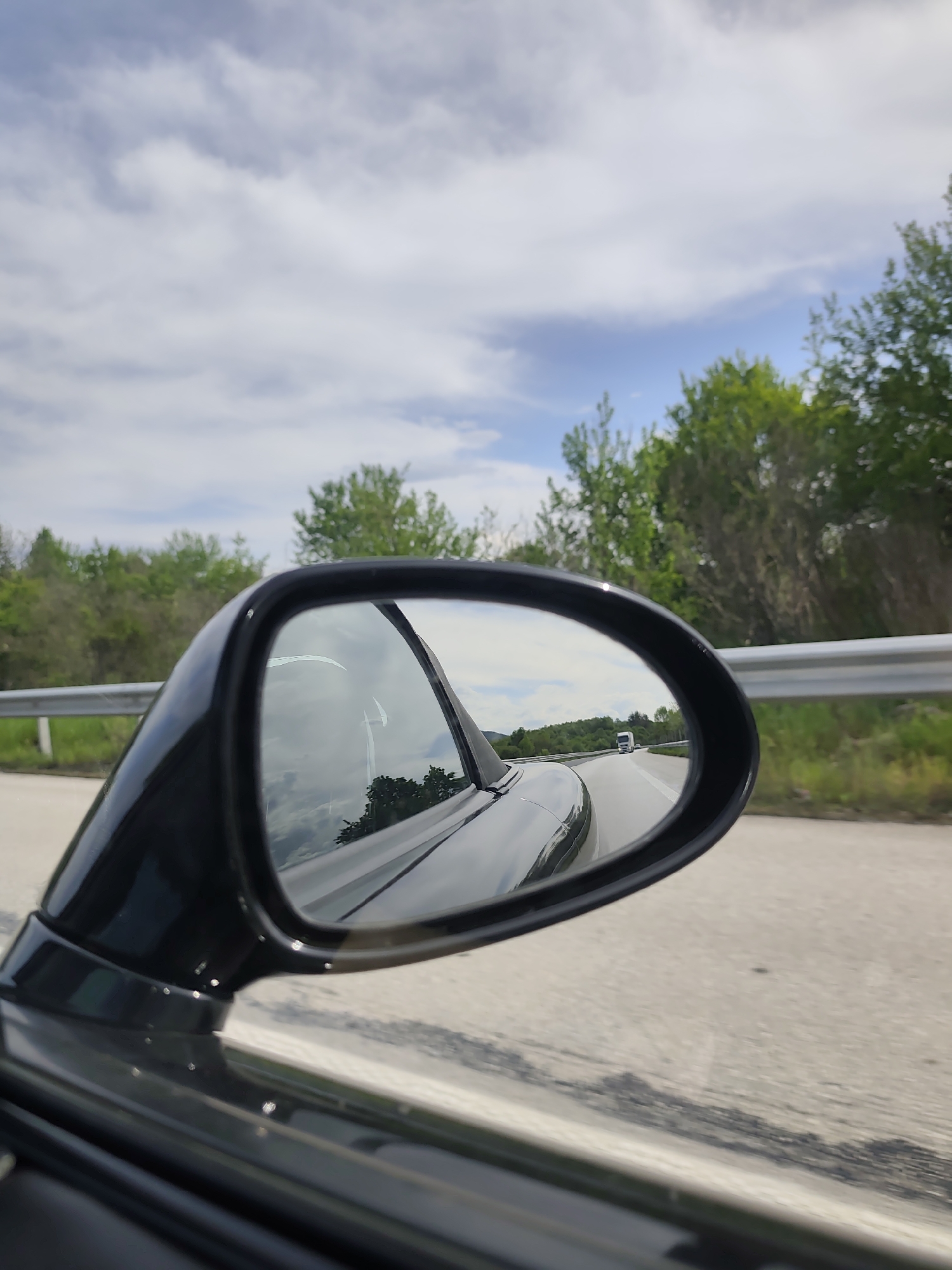 A view from a car's side mirror shows a highway scene under a sky with scattered clouds. The mirror reflects a portion of the car's body, the road ahead, and a white vehicle in the distance. A metal guardrail lines the edge of the road, and trees and greenery border the highway.