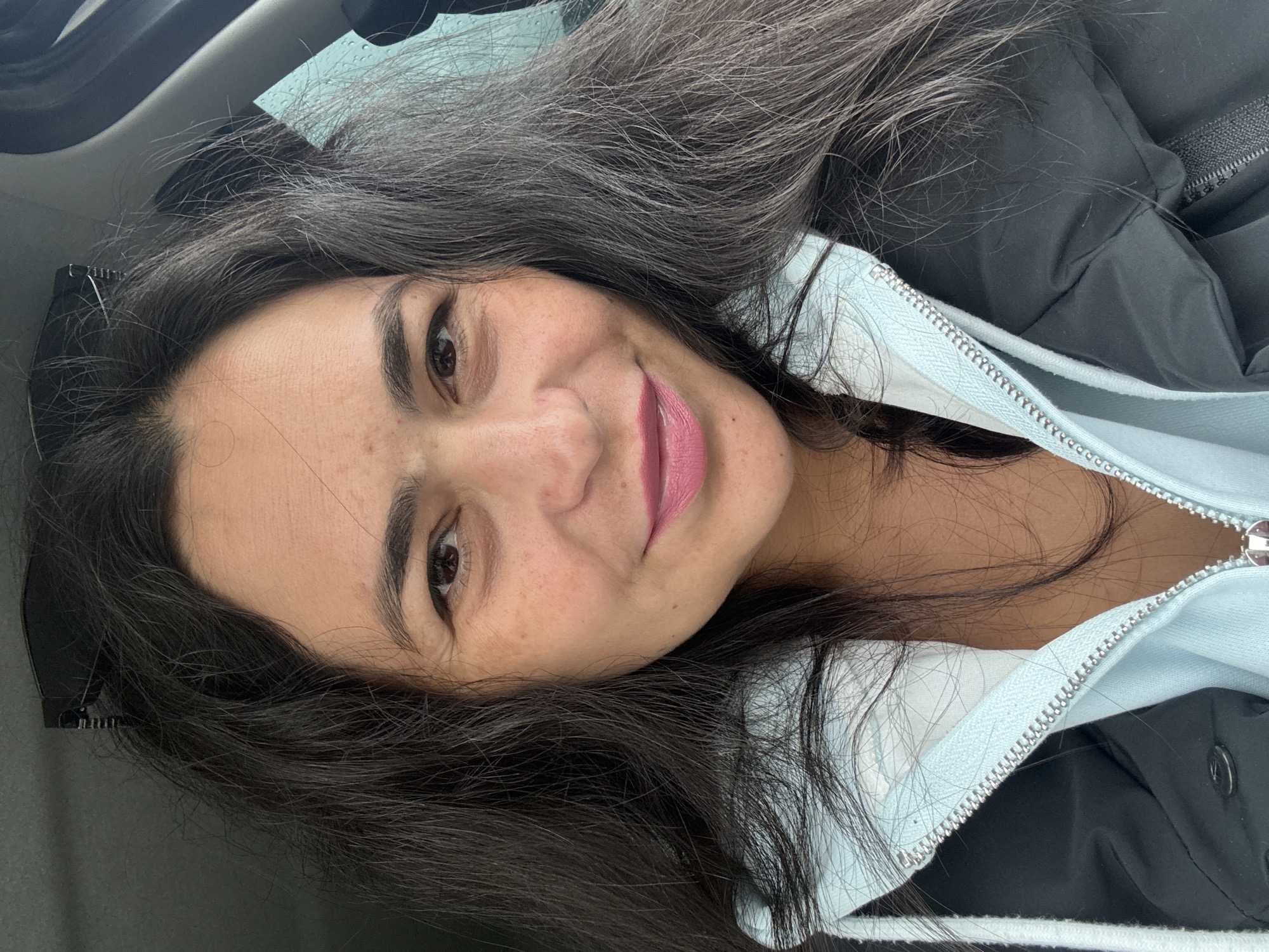 A selfie of a woman with dark hair, brown eyes, and freckles, wearing pink lipstick. She is wearing a blue and black jacket. The location cannot be determined to be Kauchuk Stantsiya, Russia, based on the visual information provided.