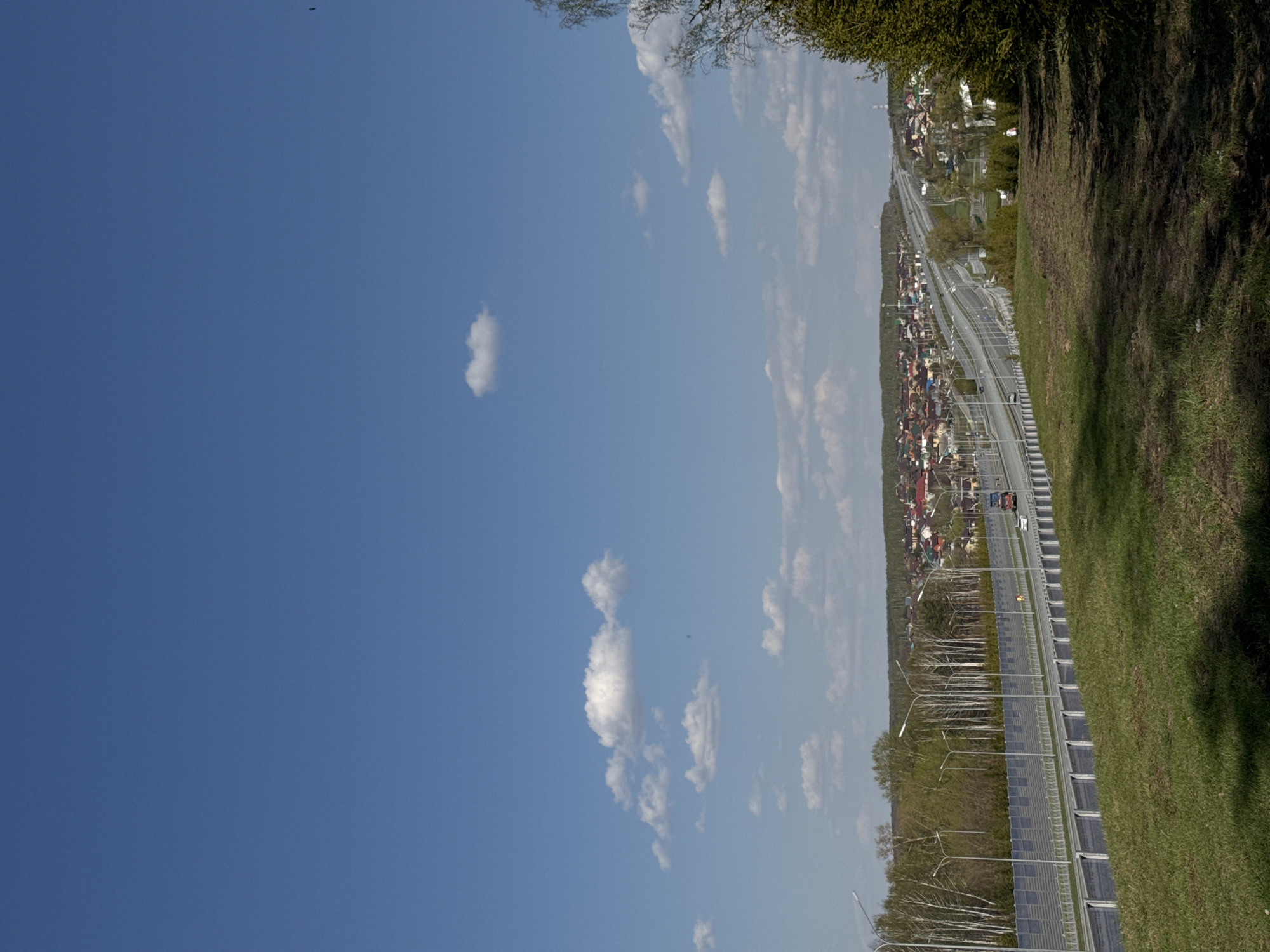 The image shows a view of a highway running alongside a residential area and a grassy embankment under a blue sky with scattered clouds. The location appears to be near Kazan, Russia, based on the architectural styles of the buildings in the residential area. The highway is lined with streetlights and what appears to be solar panels along the median. A few vehicles can be seen on the road, including what looks like a truck and some cars. The grassy embankment slopes down towards the highway, with trees and vegetation visible. The sky is predominantly blue with some light, fluffy clouds.