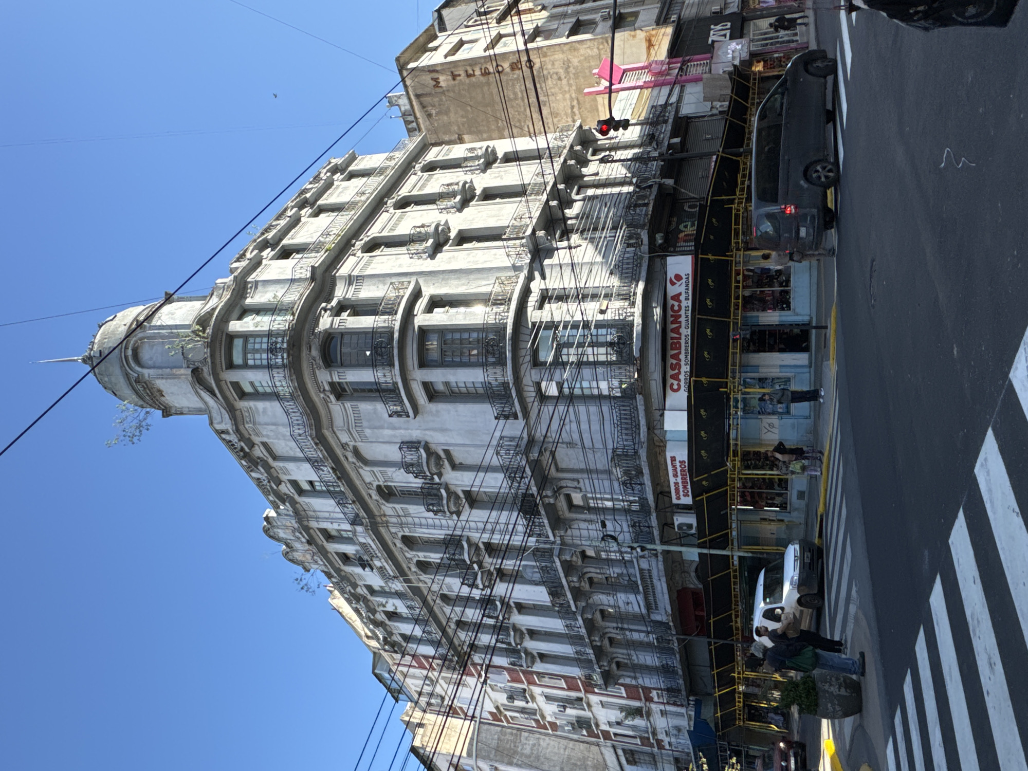 The image presents a bustling street scene in Buenos Aires, dominated by an imposing, multi-story building with ornate architecture. The building features a curved corner, elaborate balconies with railings, and a distinctive domed turret topped with a spire, suggesting a historic European architectural influence. Its light-colored facade shows signs of age and weathering, with some small plants growing from its ledges.
The scene unfolds on a clear, bright day under a vivid blue sky, with numerous black utility wires crisscrossing above the street. At ground level, a white pedestrian crosswalk stretches across the asphalt. Several vehicles are visible, including a dark grey van parked near the curb and a white car further down the street. A few pedestrians are seen walking on the sidewalk and near the crosswalk, indicating daily urban activity.
On the building's ground floor, a shop storefront is prominent, displaying a red and white sign that clearly reads "CASABLANCA." Below this, smaller text translates to "Hats - Hats - Gloves - Scarves" ("GORROS - SOMBREROS - GUANTES - BUFANDAS"). Yellow scaffolding is erected along the lower part of the main building, possibly for maintenance or renovation. A red traffic light is visible suspended over the street. Another building in the background features partially visible text, "MARUANO." The image captures a vibrant snapshot of city life, blending grand old architecture with contemporary street activity.