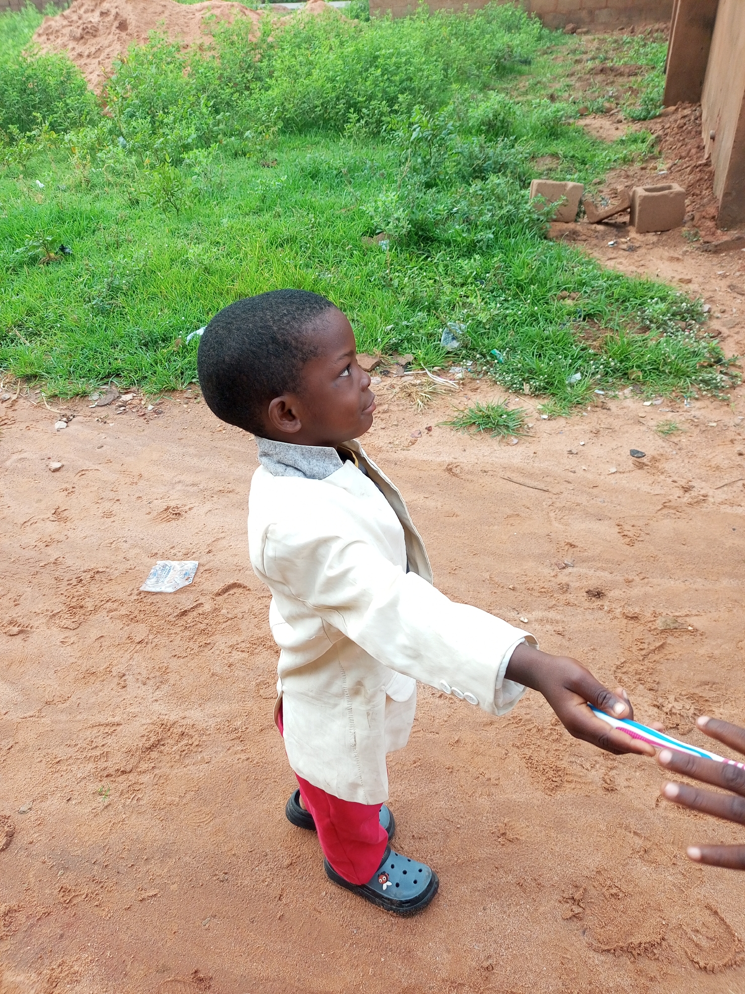 A young Black boy stands outdoors on a dirt and sand surface, looking to his right. He is wearing a cream-colored blazer over a gray collared shirt and red pants. His feet are clad in gray Crocs with a red and white cartoon character on them. The boy's right hand is extended, holding a pink and blue striped toothbrush which he is passing to another person whose hands are visible on the right side of the frame.
The environment is characterized by dry, reddish-brown earth with scattered small rocks and footprints. In the background, there is a patch of green grass and bushes. To the far right, a portion of a brown brick wall is visible. The scene appears to be a simple, outdoor setting, possibly a residential area or a communal space in Tempure, Nigeria. The lighting suggests it is daytime.