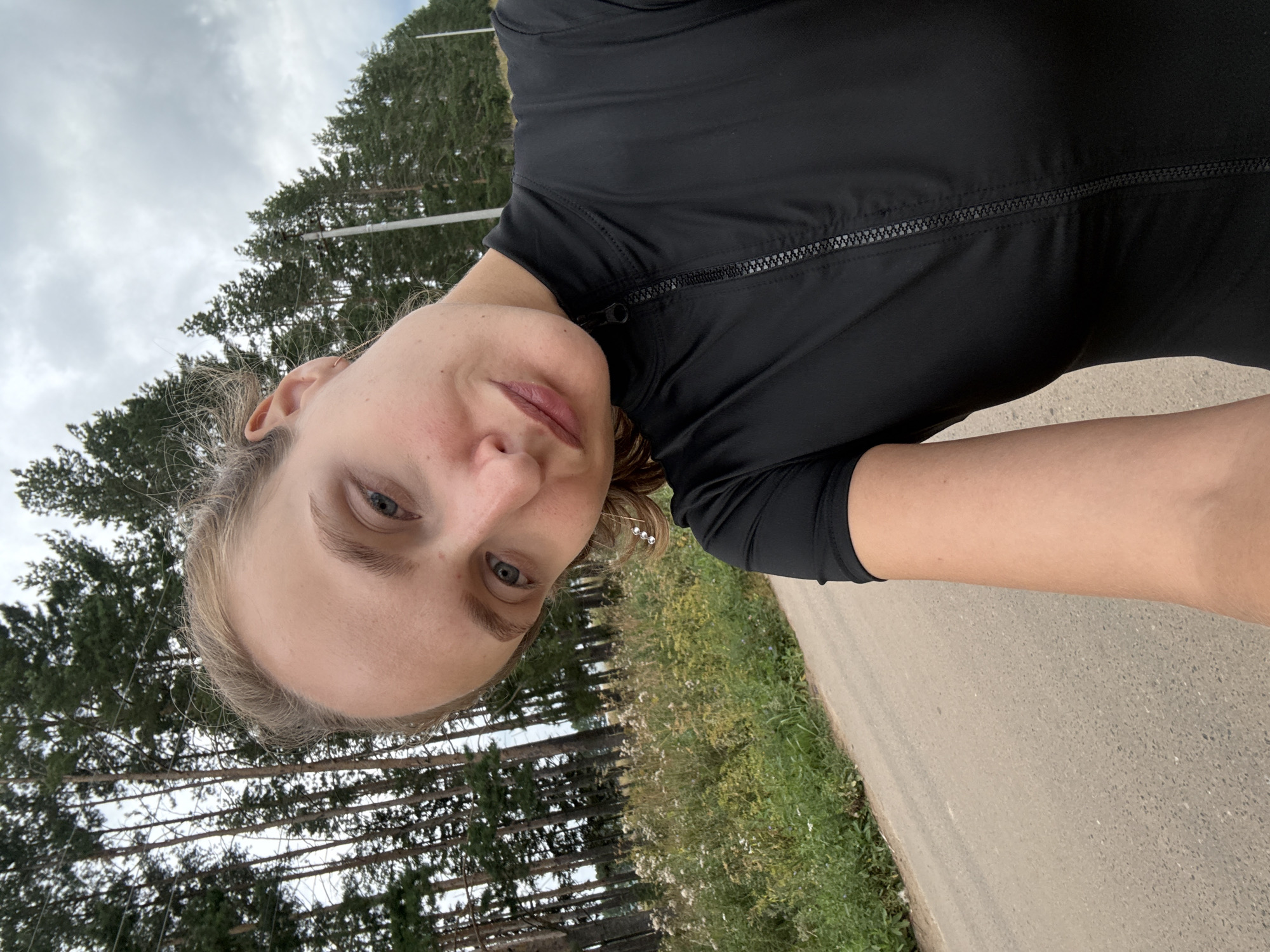 This is a selfie of a young woman outdoors. She is wearing a black zip-up top. The background consists of a road, grassy vegetation with wildflowers, and a dense forest of pine trees under an overcast sky. The photo appears to be taken in the late afternoon. The woman is looking at the camera with a neutral expression. There are no visible text elements in the image. The orientation of the image is rotated 90 degrees counter-clockwise.