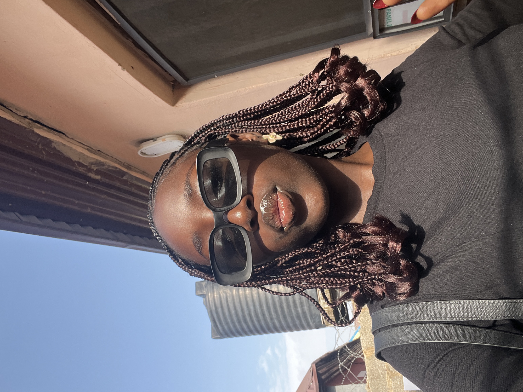 A medium shot depicts a woman wearing sunglasses, turned sideways. The woman has dark skin and is wearing box braids. She is also wearing a black shirt and a handbag. A silver water tank is partially visible. The sky is pale blue. The photo was taken outdoors in Abuja, Nigeria. The exact location is indeterminable.