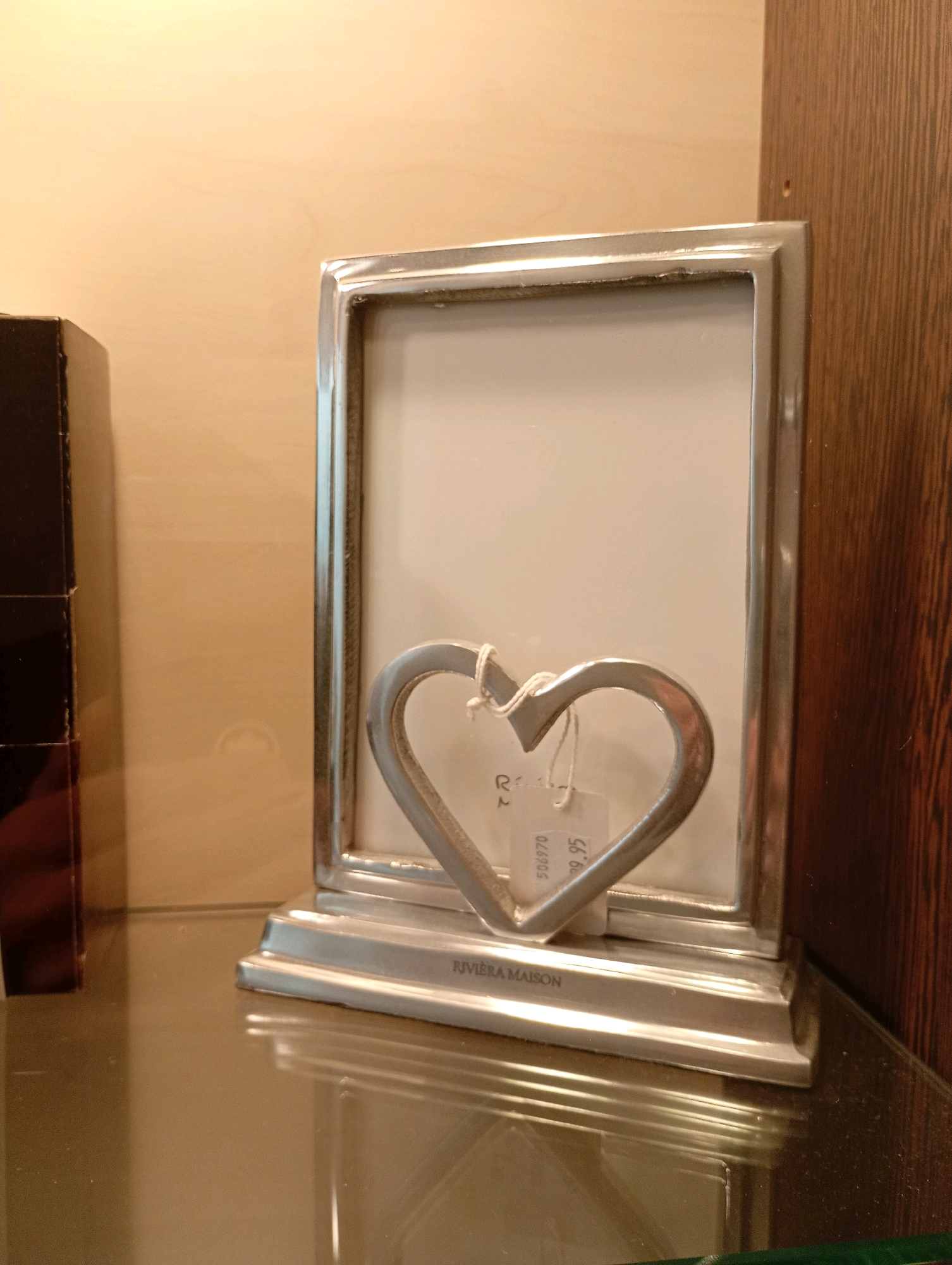 The image shows a silver-toned, rectangular picture frame with a decorative heart-shaped ornament hanging in front of it. The frame is standing upright on a reflective surface, likely a glass shelf, which is placed against a wooden panel with vertical grain. To the left of the frame, a dark, rectangular object is partially visible. A small white tag with text and numbers is attached to the heart ornament with white string. The background wall is a light, neutral color. The scene appears to be an indoor setting, possibly a display or a home environment. No people are present in the image. The location is not specified in the image.