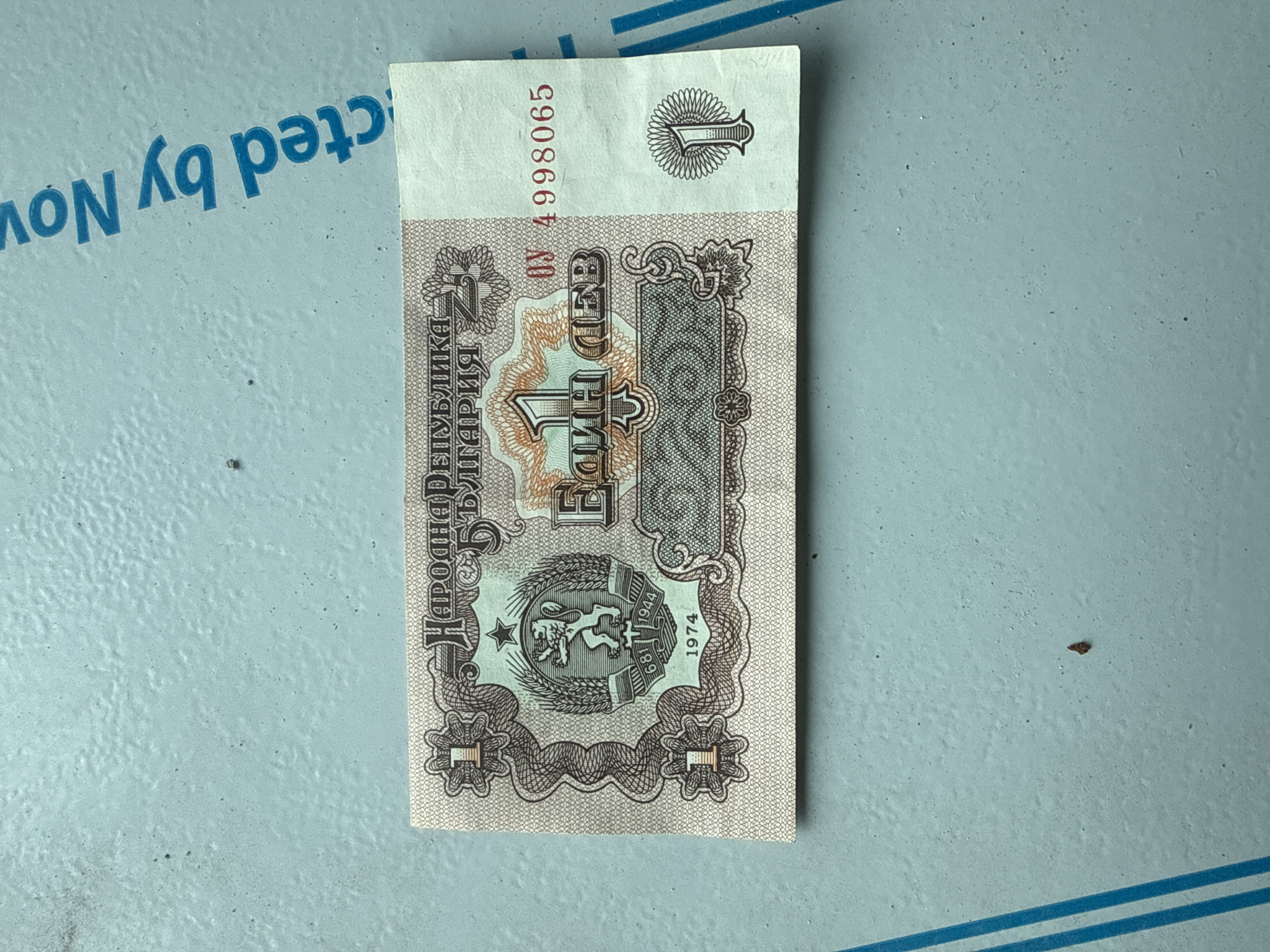The image shows a 1 Bulgarian lev banknote from 1974. The banknote features the Bulgarian coat of arms with a lion, the text "Народна Република България" (People's Republic of Bulgaria), and the denomination "Един Лев" (One Lev). The serial number "ΟΥ 4998065" is printed on the top right. The banknote is resting on a light blue surface. There is no information to suggest the specific location of the object is in Sofia, Bulgaria.