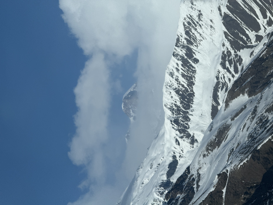 The scene depicts a snow-covered mountainous landscape, identified as being located in the city of Kanobi, Georgia. The upper third of the image consists of a clear, deep blue sky. Below this, a band of white cumulus clouds partially obscures the horizon. Through a gap in these clouds, a distant, prominent, rugged peak, heavily coated in snow, is visible, flanked by a lesser, partially cloud-enshrouded peak to its left. The mid-ground and foreground are dominated by multiple layers of snow-covered mountain ridges and slopes. The upper mid-ground mountains exhibit significant snow accumulation interspersed with exposed dark rock formations, particularly on steep inclines, creating a striped or patterned effect. The lower foreground slopes show a less continuous snow cover, revealing patches of dark, possibly rocky or barren earth between snowdrifts. No individuals, man-made structures, or signs of activity are present within the depicted environment.