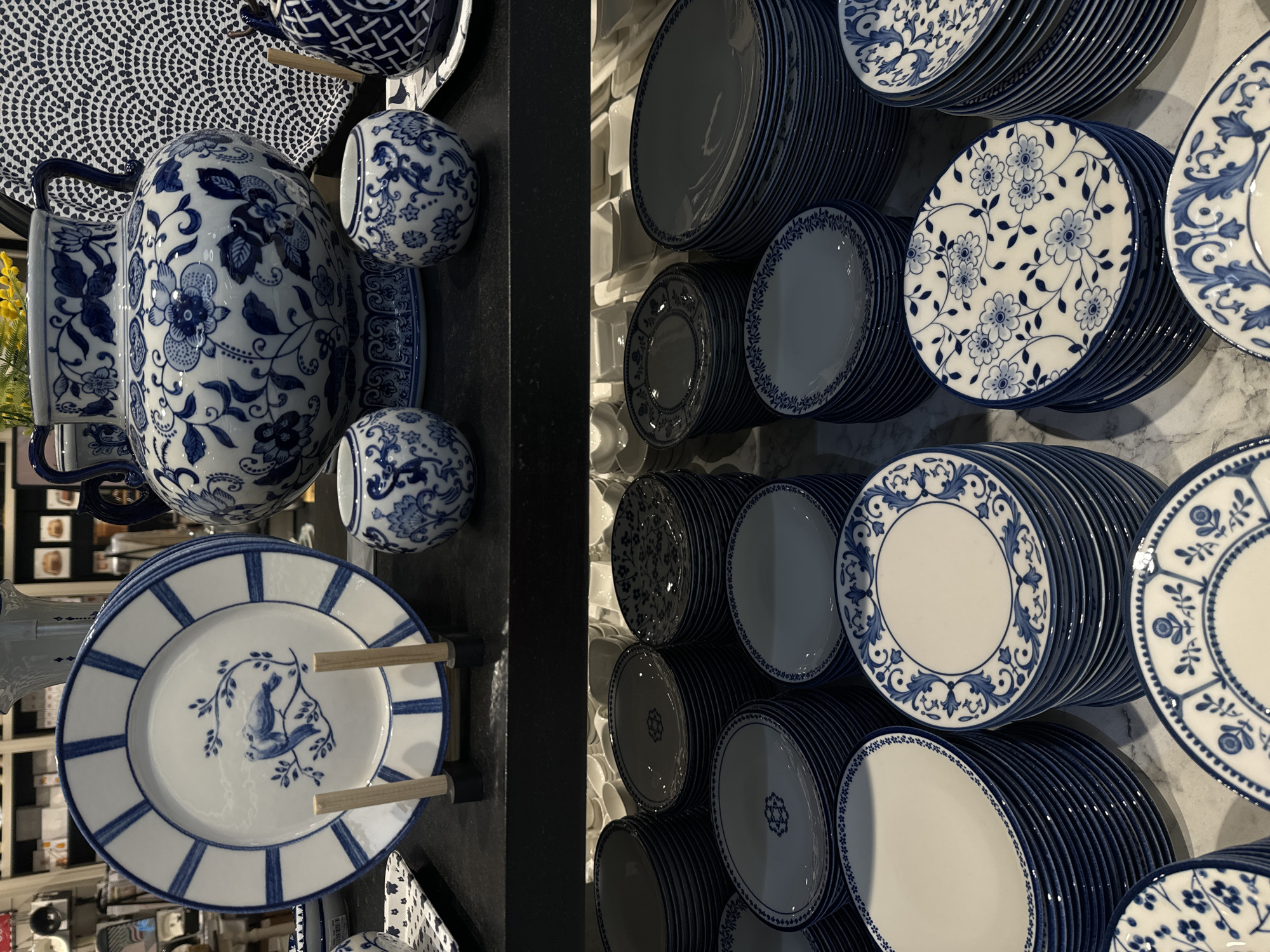 The image shows a display of blue and white porcelain tableware. On the left, a black shelf holds a large floral-patterned pitcher, two small matching cups, and a plate with a blue bird design. To the right, stacks of plates in various blue and white patterns are arranged on a light-colored wall. The setting appears to be a retail environment, possibly a store in Yudino, Russia.