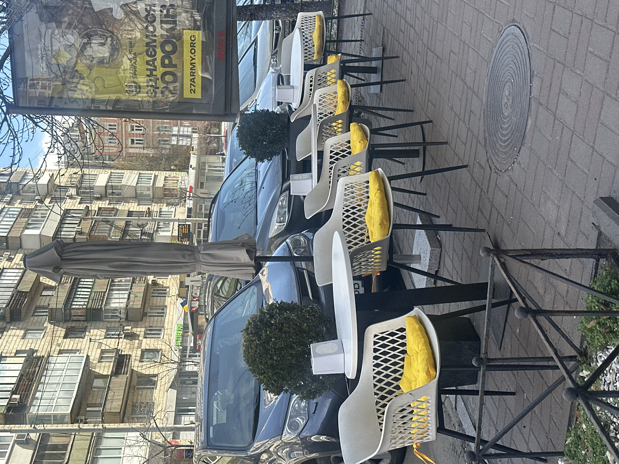 The image shows an outdoor cafe scene in Kyiv, Ukraine. Several white plastic chairs with yellow cushions are arranged alongside small tables. Cars are parked closely along the street adjacent to the cafe seating. A large advertisement is positioned prominently on the left side of the image, with text including "27 ARMY ORG." Buildings with multiple apartments are visible in the background. The scene is situated on a paved sidewalk with a manhole cover visible on the ground.