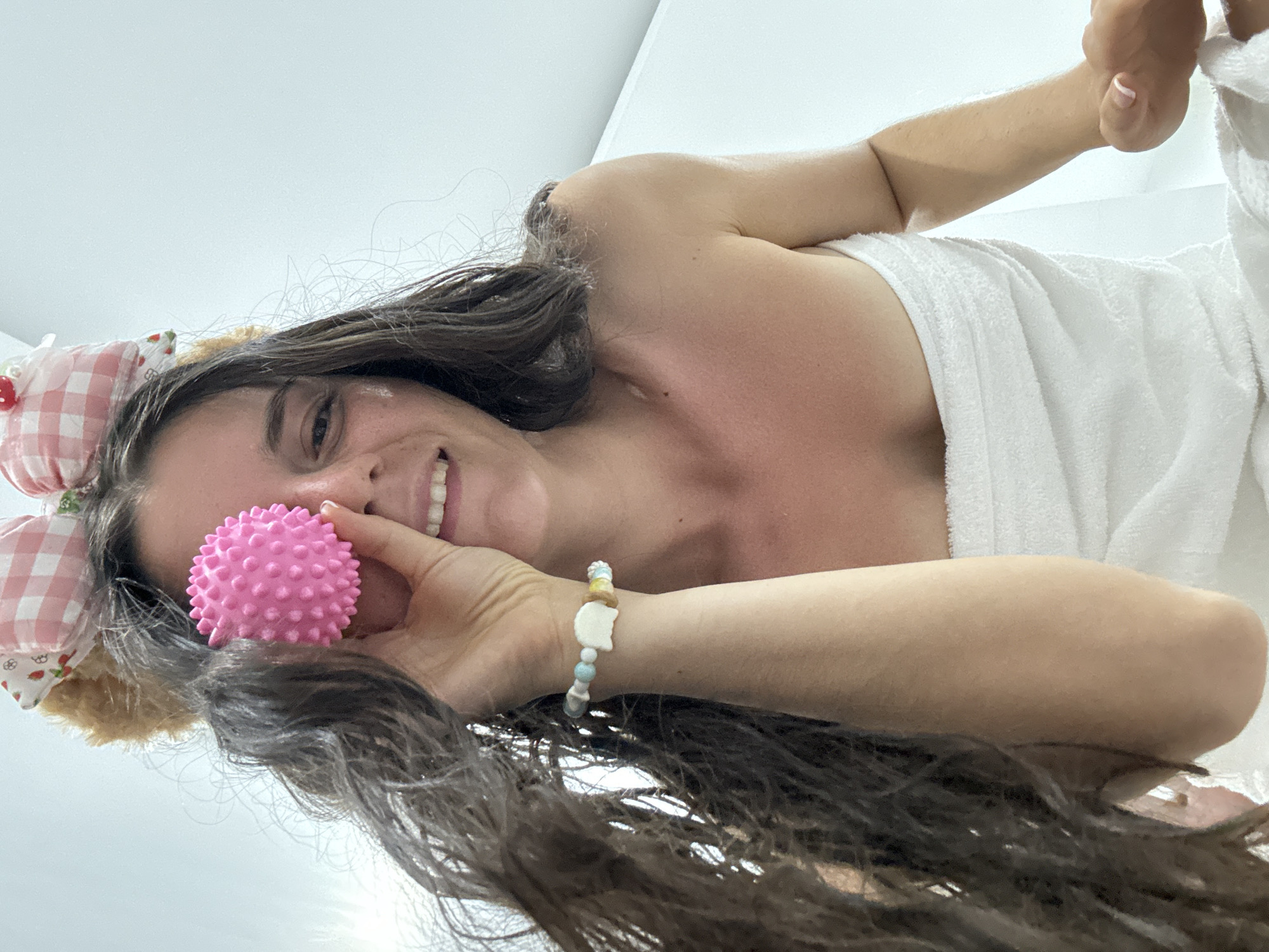 A smiling woman in a white towel is lying down, holding a pink spiky ball up to her face. She is wearing a headband with bunny ears. Her hair is dark brown and wavy. She is also wearing a beaded bracelet on her left wrist. The location appears to be an indoor bathroom with white walls. It is impossible to determine the location is Mũi Né, Vietnam from just the provided image.