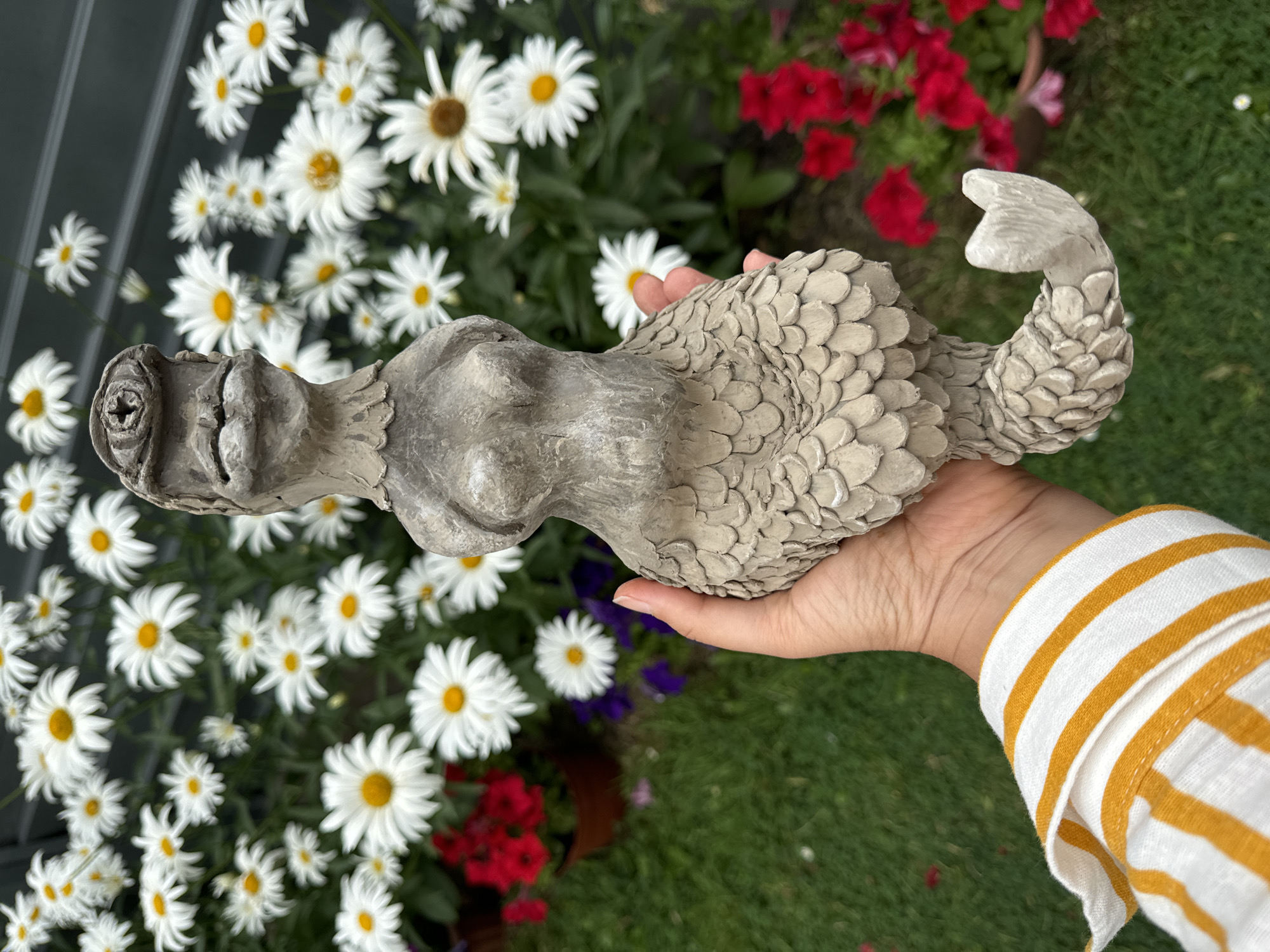 A person's hand, wearing a white and yellow striped shirt sleeve, holds a grey, concrete-like sculpture of a mermaid. The sculpture has a human-like torso with breasts, and a scaled lower body that tapers into a fishtail. The mermaid's head is somewhat grotesque, with a beak-like mouth and a peculiar eye socket. Behind the sculpture and hand, a lush garden is visible, filled with numerous white daisies with yellow centers, and some red and purple flowers in pots and the ground. The background also includes greenery and a dark, greyish structure, possibly a fence or wall. The scene appears to be outdoors.